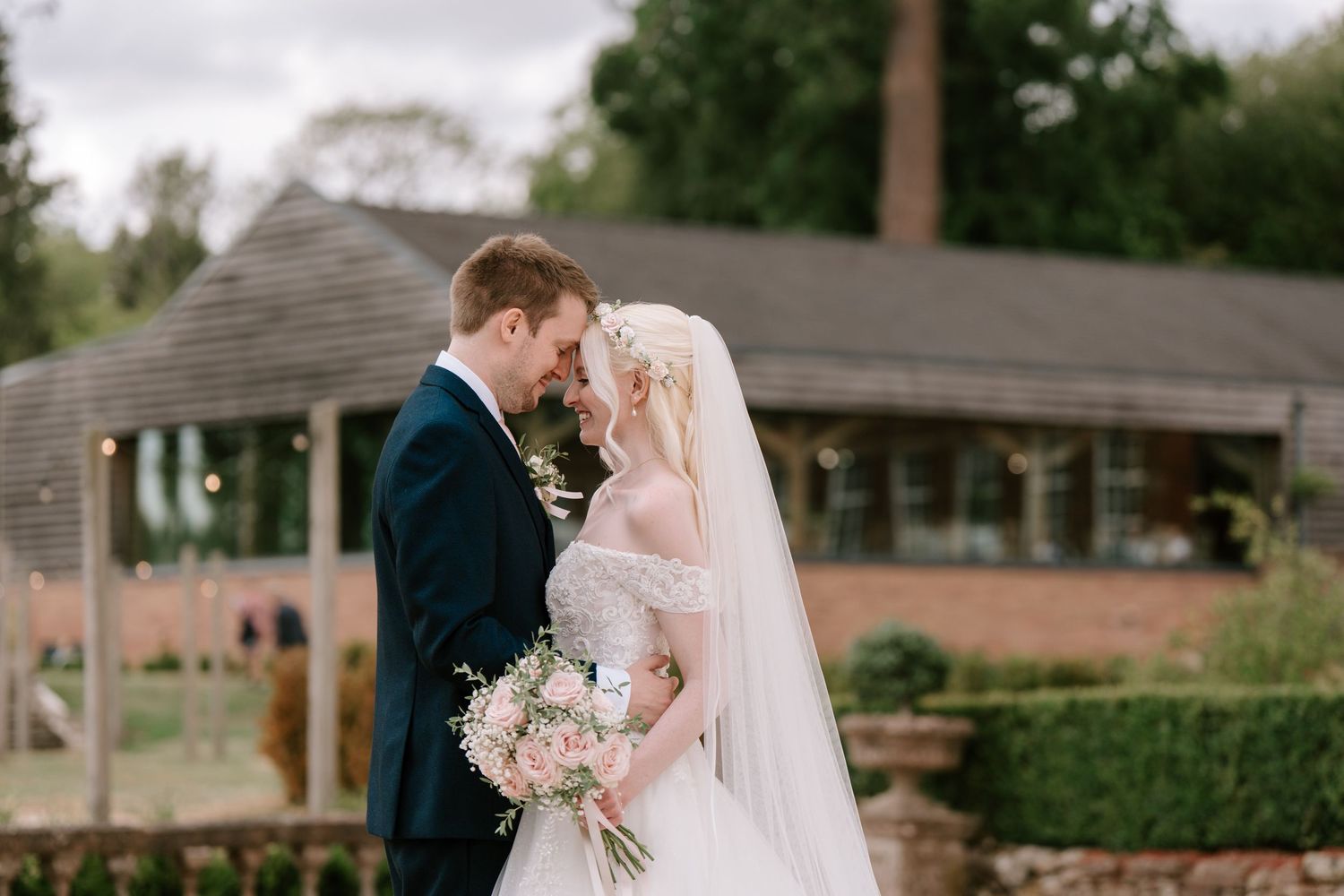 Bride and groom share a romantic moment in a garden, bride holding a pink bouquet in a white gown with veil.