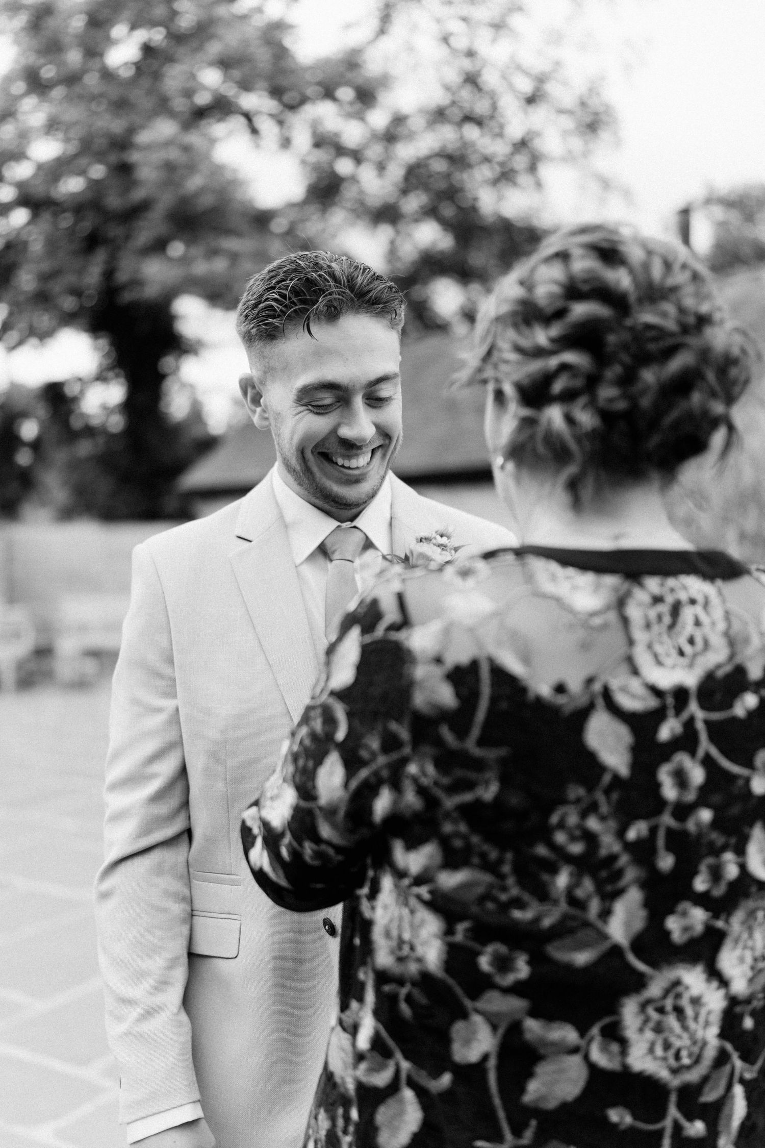 Black and white photo of a smiling groom in a light suit sharing a tender moment with a woman in a floral dress outdoors.