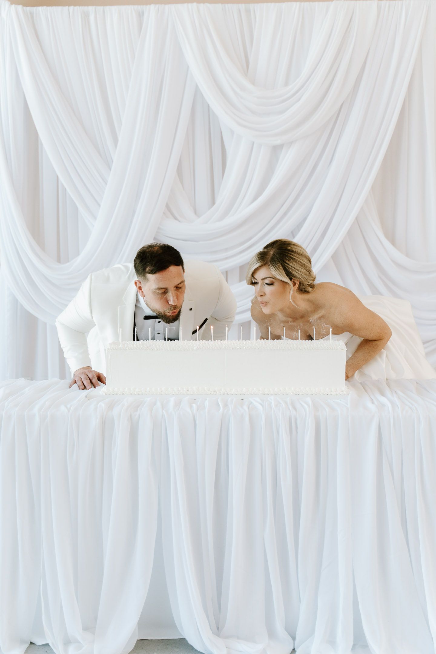 Bride and groom blowing out candles on white wedding cake at elegant reception with white draped backdrop.