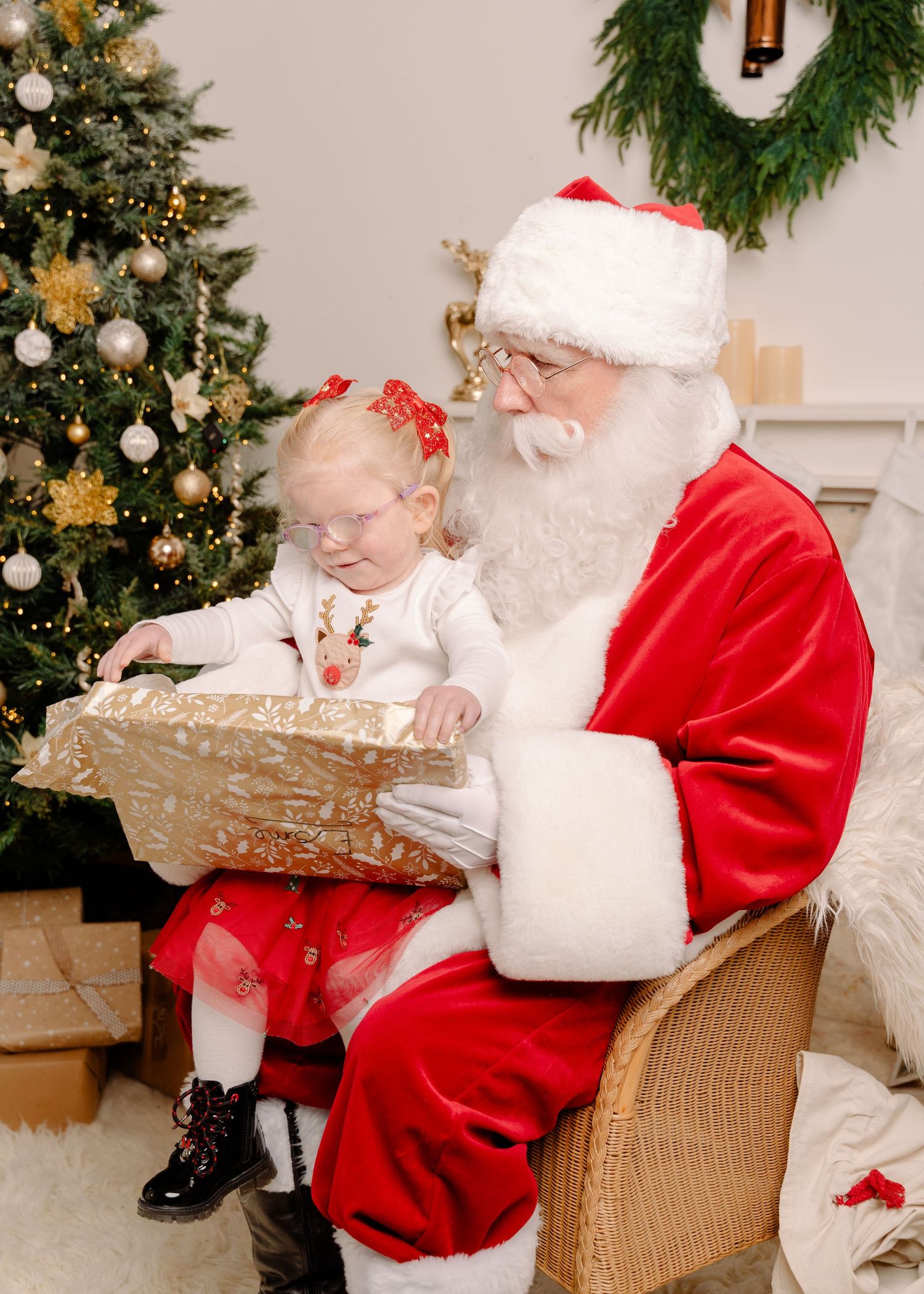 A baby girl opens a gold wrapped gift while sitting on Santa Claus lap beside a Christmas tree.