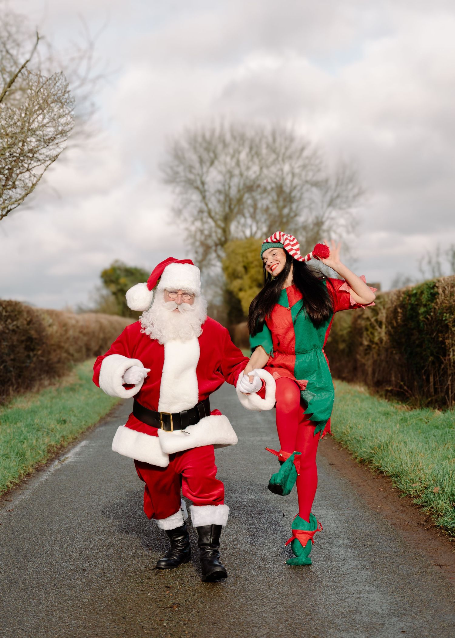 Santa Claus and a festive elf dancing joyfully on a rural road surrounded by hedgerows and trees.