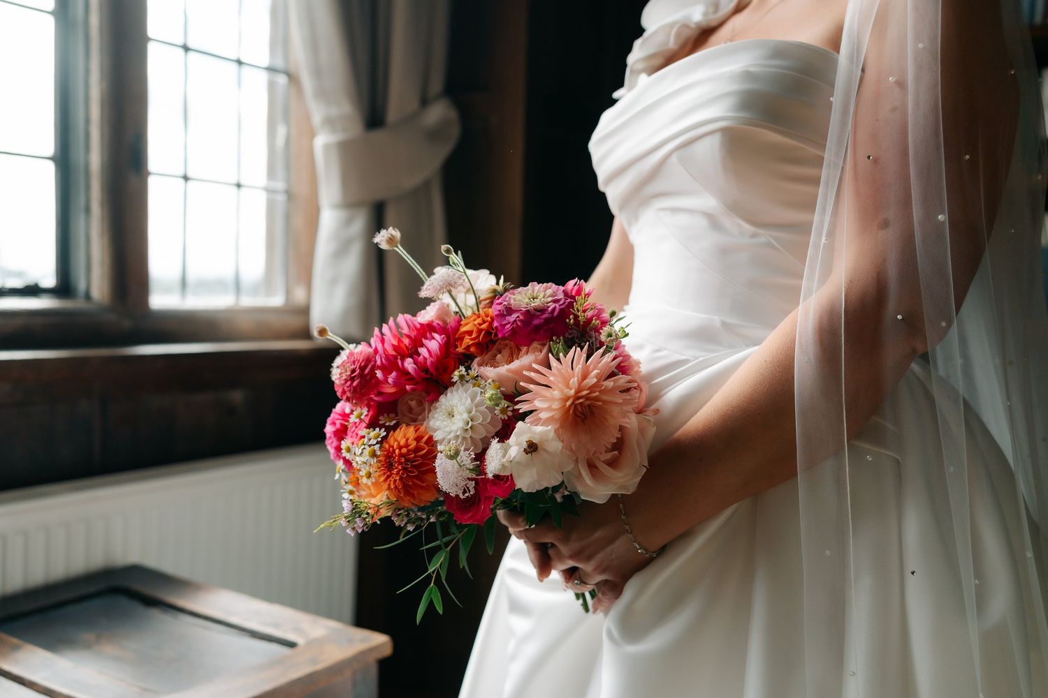Bride in white strapless gown holding vibrant pink and orange floral bouquet with veil near rustic window.