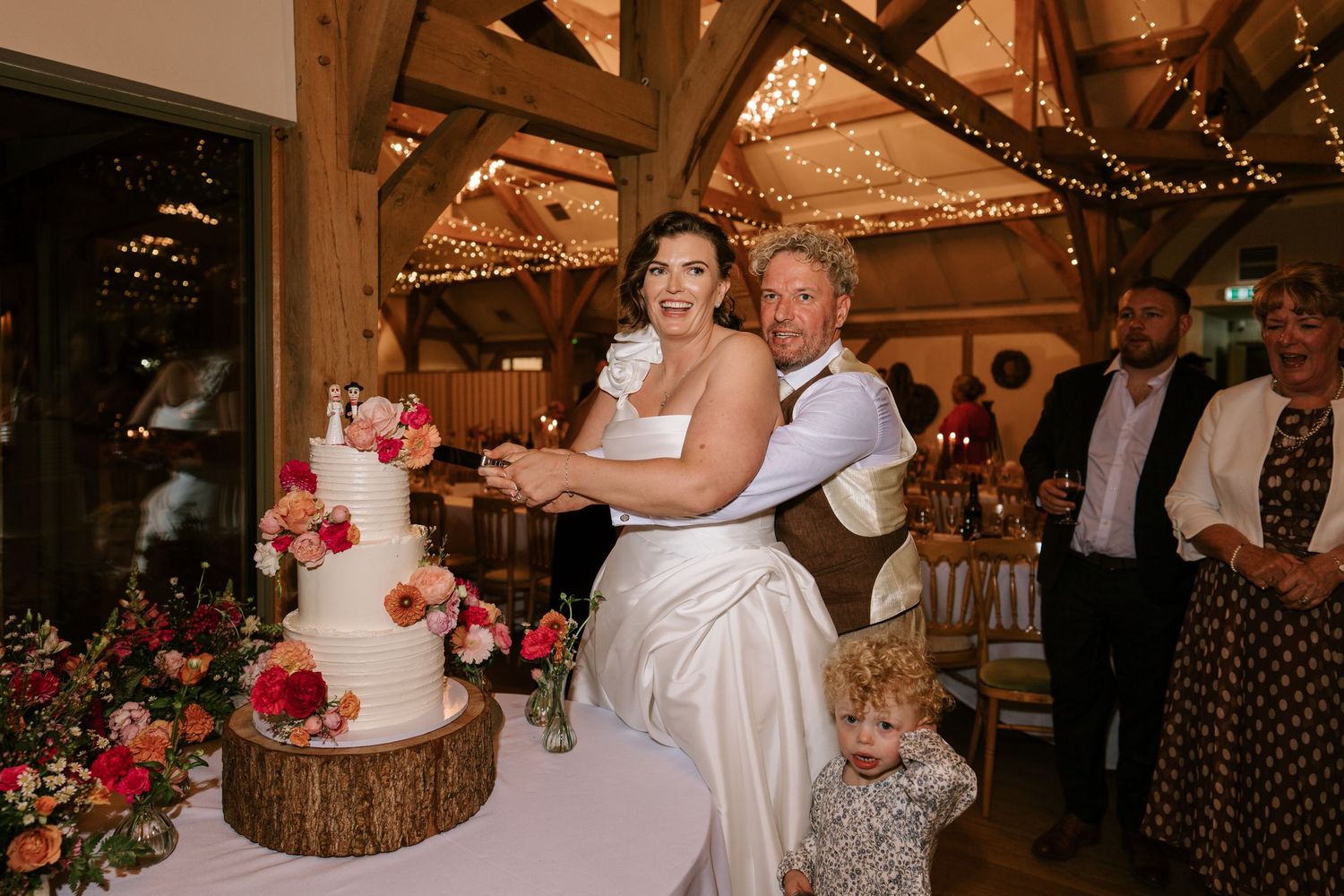 Bride and groom cutting tiered wedding cake decorated with flowers in a rustic barn venue with fairy lights.