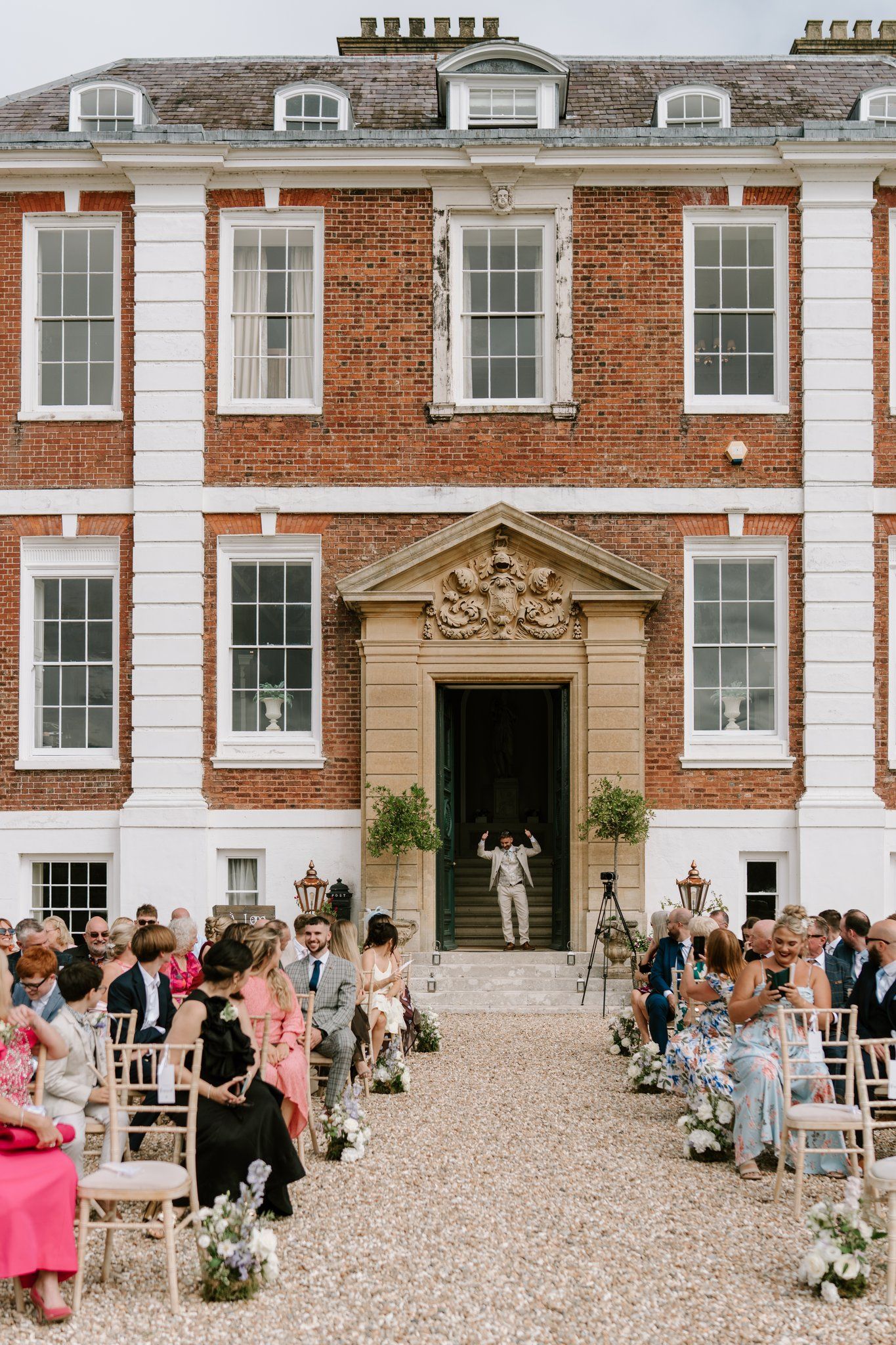 Outdoor wedding ceremony at a grand red brick Georgian manor house with guests seated along a gravel aisle.