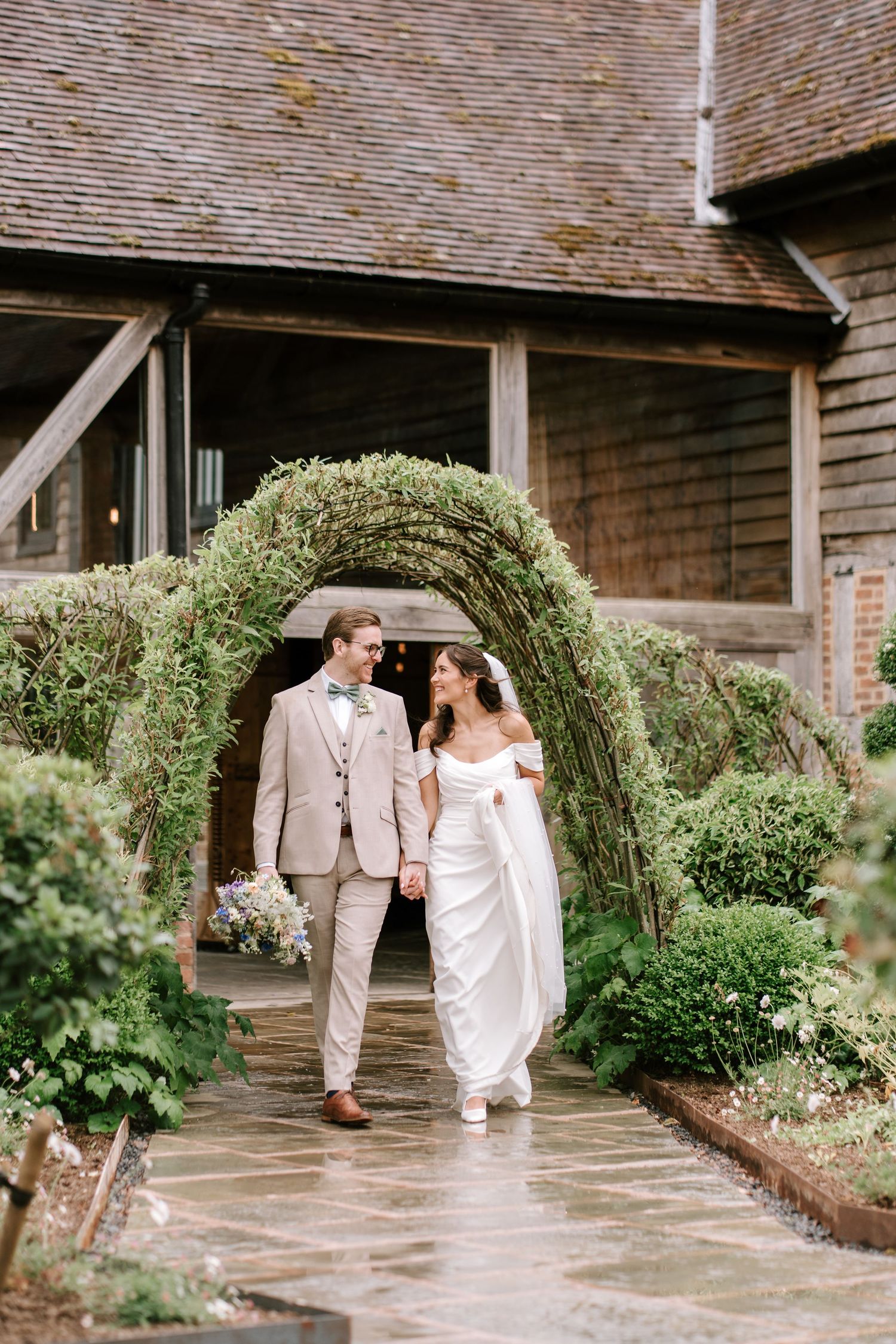 Bride and groom walk hand-in-hand through a lush green arch at a rustic barn wedding venue.