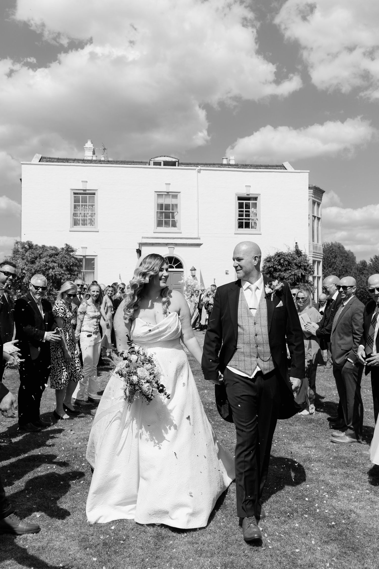 Black and white photo of a bride and groom walking through confetti at an outdoor wedding ceremony.