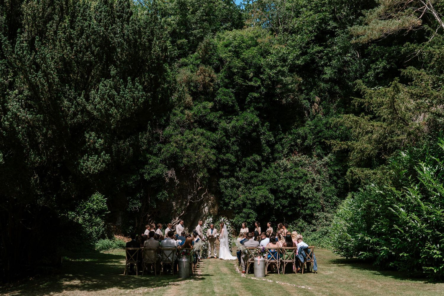 Small outdoor wedding ceremony with guests seated on chairs surrounded by lush green trees on a sunny day.