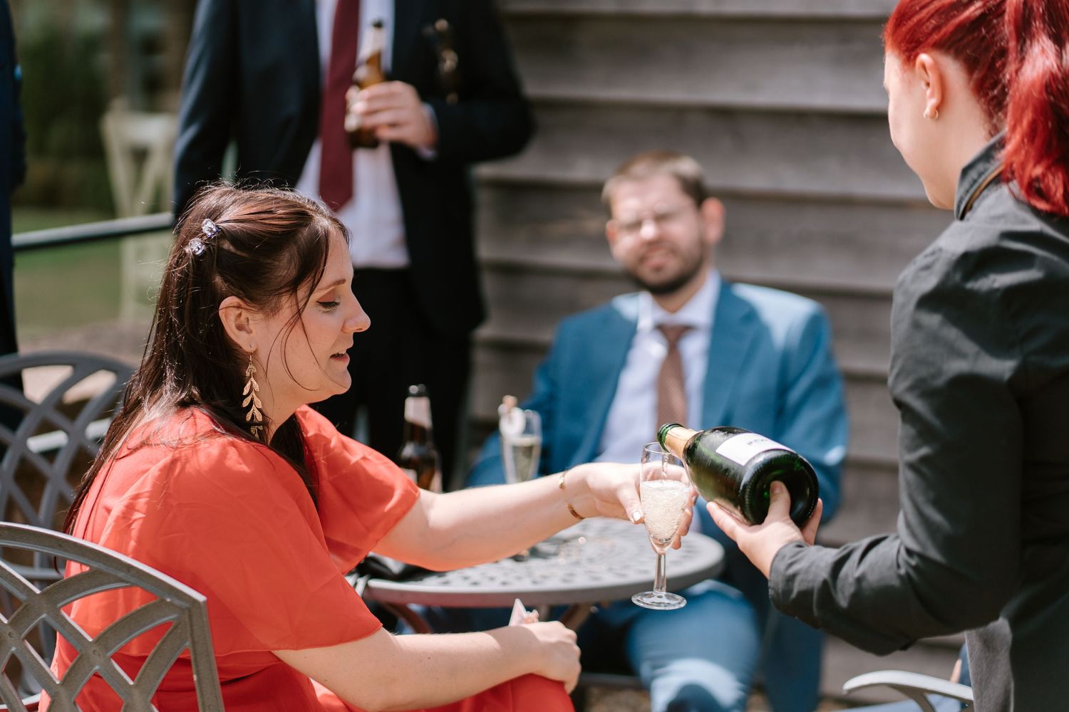 A waitress pours champagne for a woman in a red dress at an outdoor wedding reception.