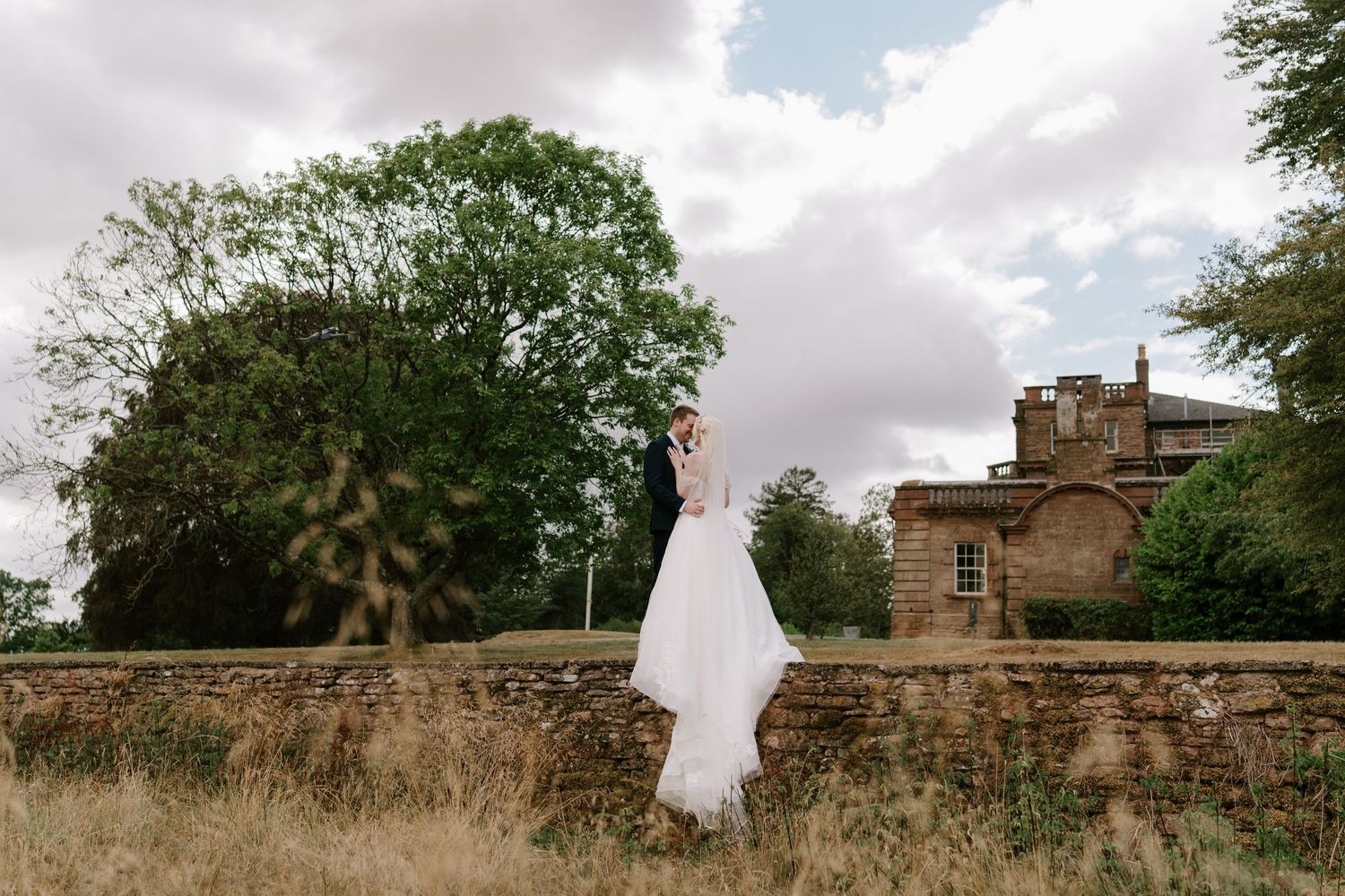 Bride and groom embrace on a stone wall at a historic estate, with lush trees and cloudy sky backdrop.