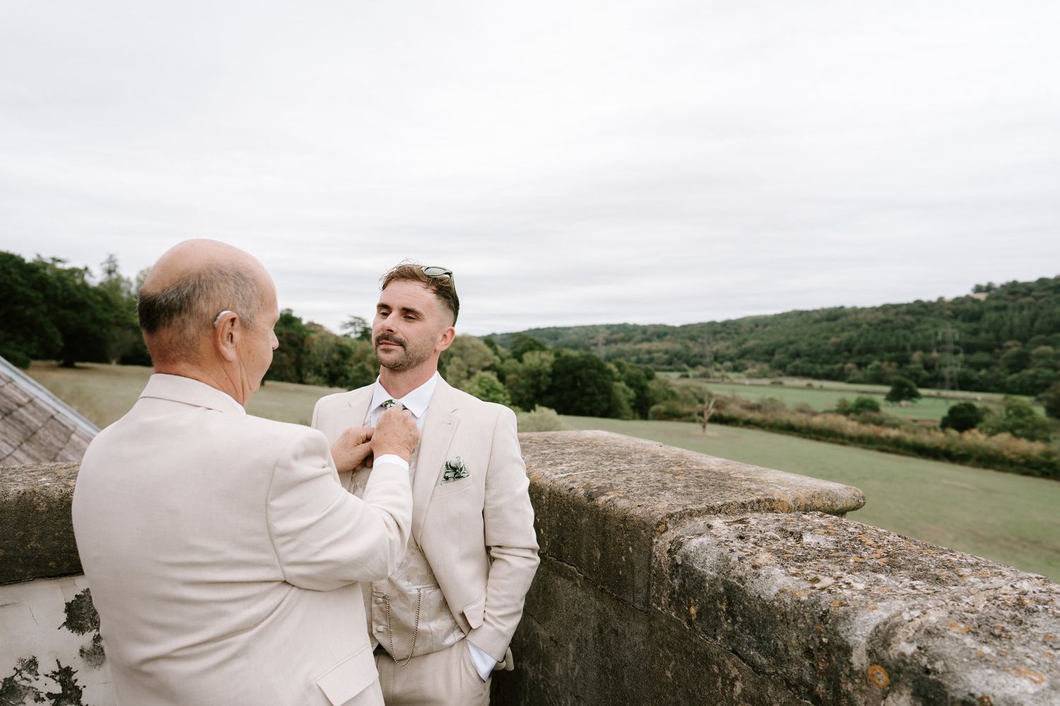 Groom in linen suit gets tie adjusted by older man on stone terrace with lush green countryside backdrop.