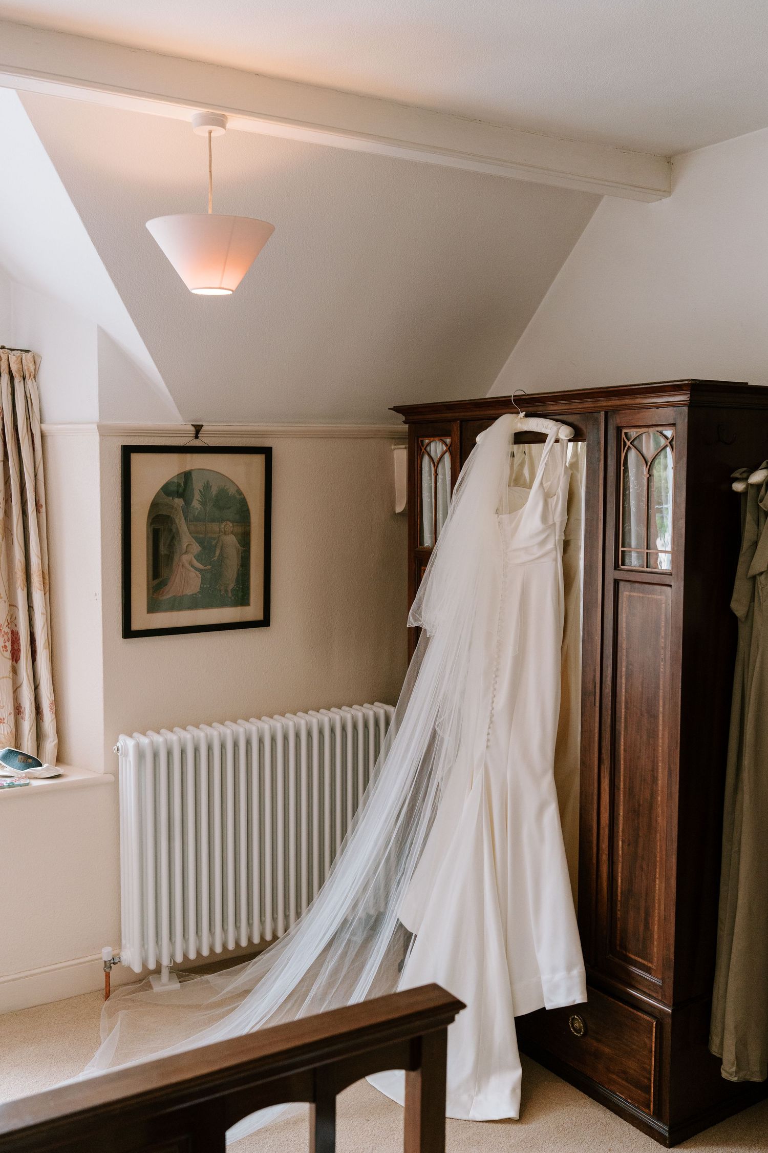 White wedding dress with veil hanging on dark wooden wardrobe in elegant room with white radiator.