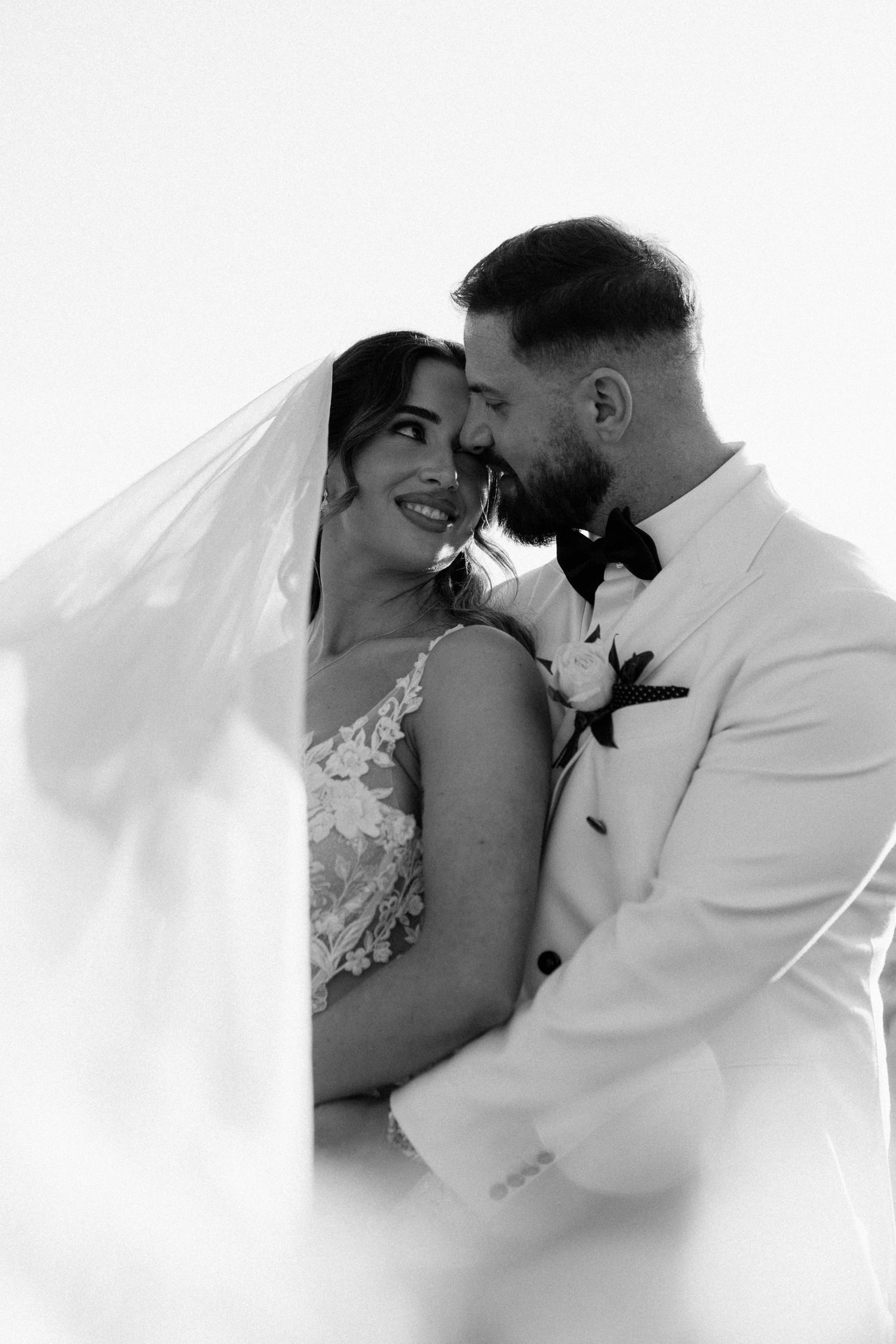 Black and white wedding photo of smiling bride and bearded groom in white suit, veil flowing around them.