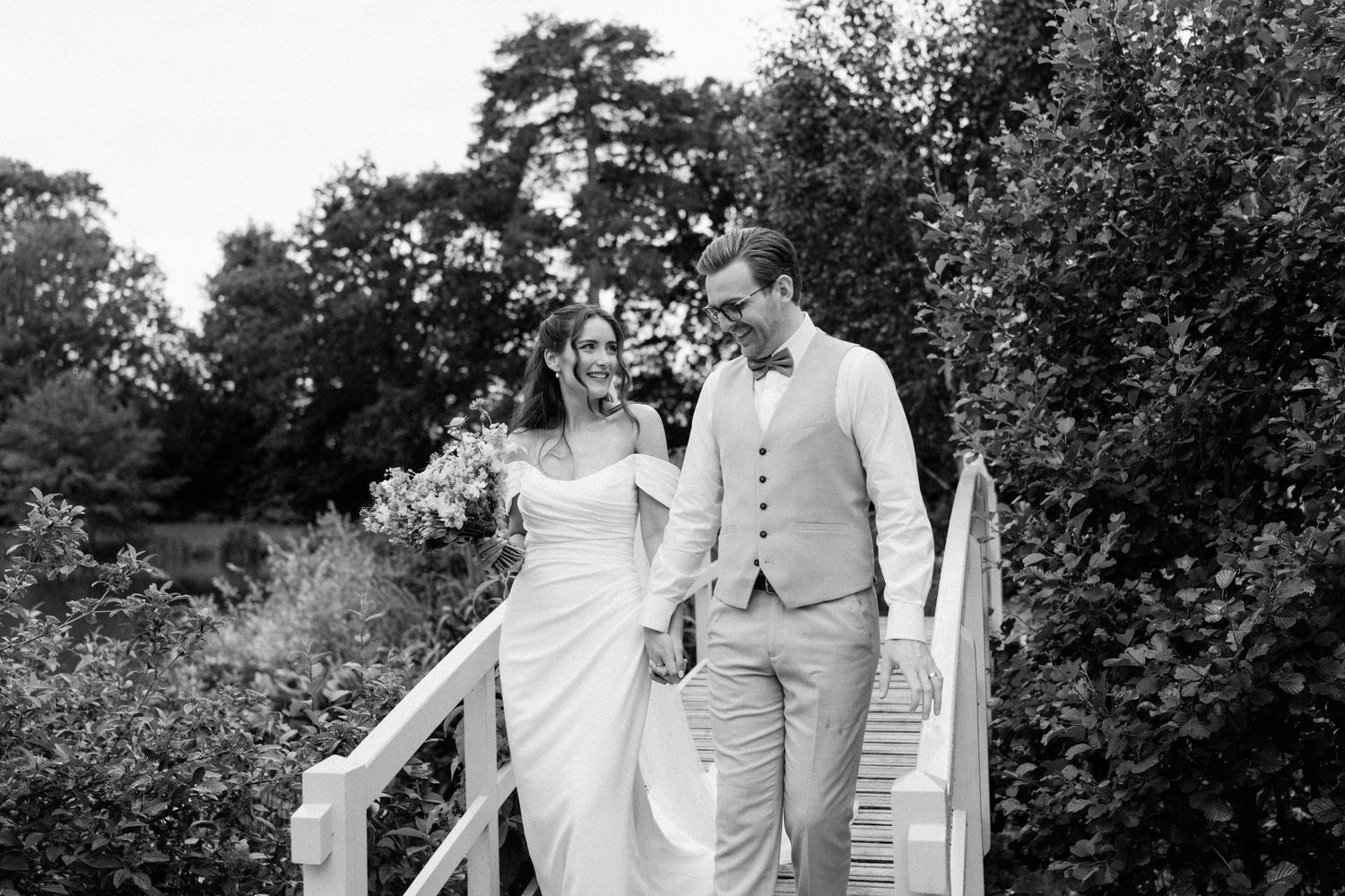 Bride and groom walking hand in hand on white bridge, black and white romantic wedding portrait.