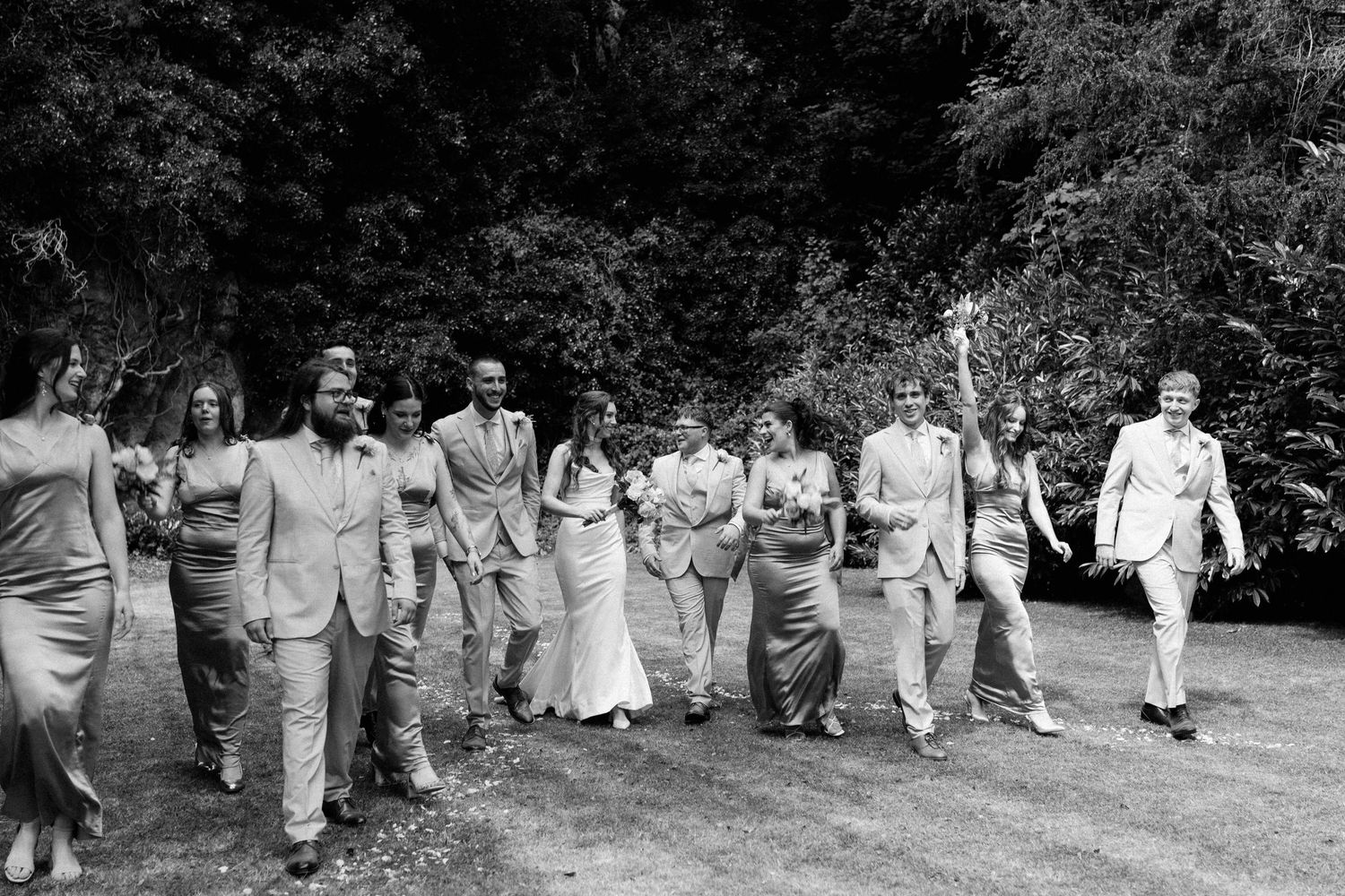 Wedding party walking together outdoors in black and white photo, bridesmaids in gowns and groomsmen in suits.