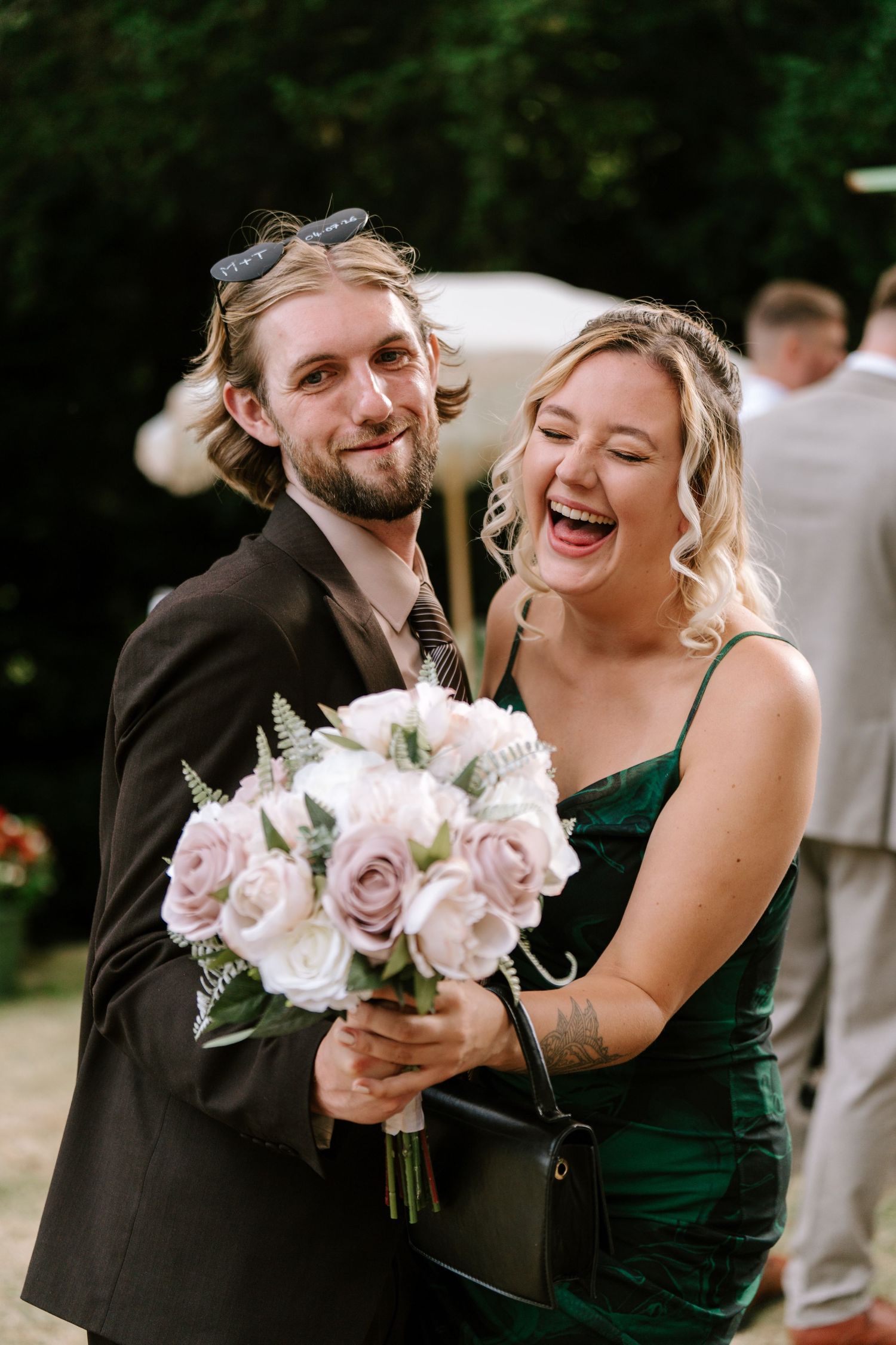 Laughing couple at a wedding holding a pink rose bouquet, man in dark suit and woman in green dress.
