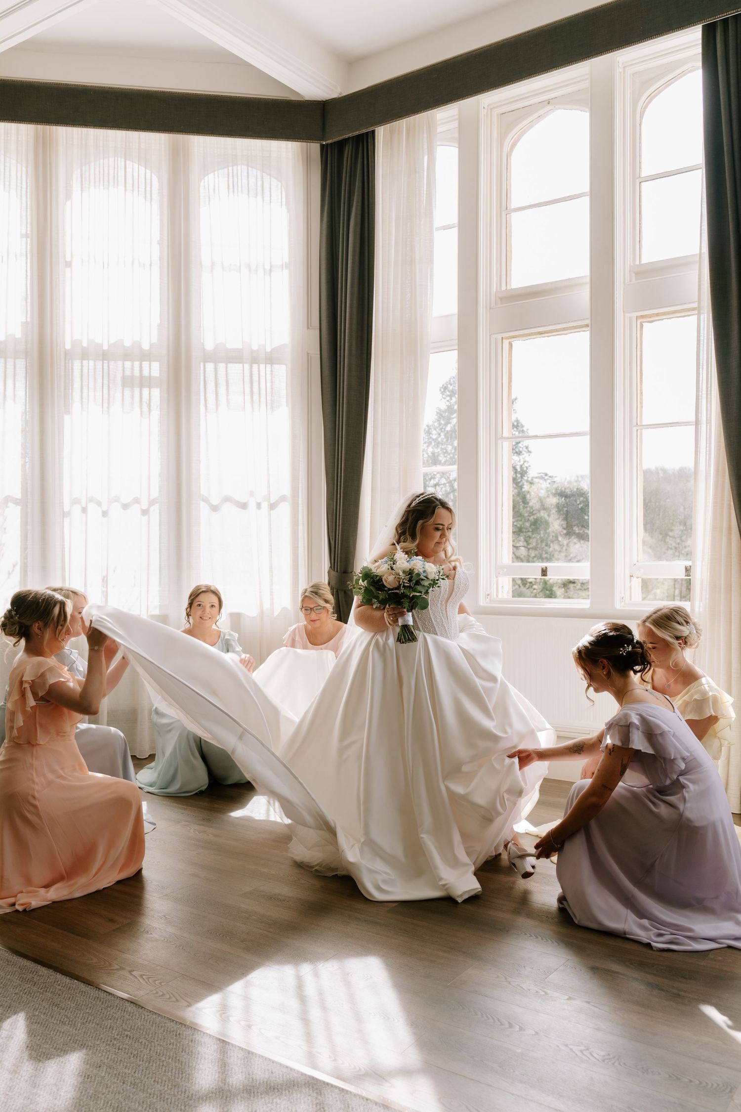 Bride in white gown holds bouquet while bridesmaids in pastel dresses adjust her dress near large windows.