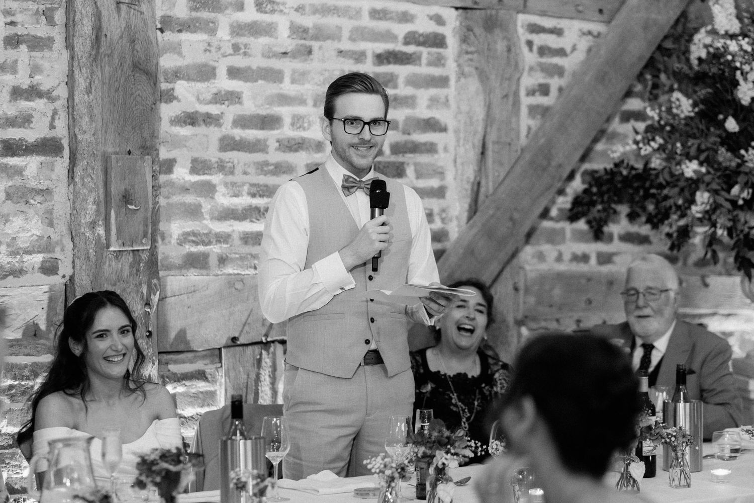 Groom delivering a speech at a rustic barn wedding reception, guests laughing, black and white photo.