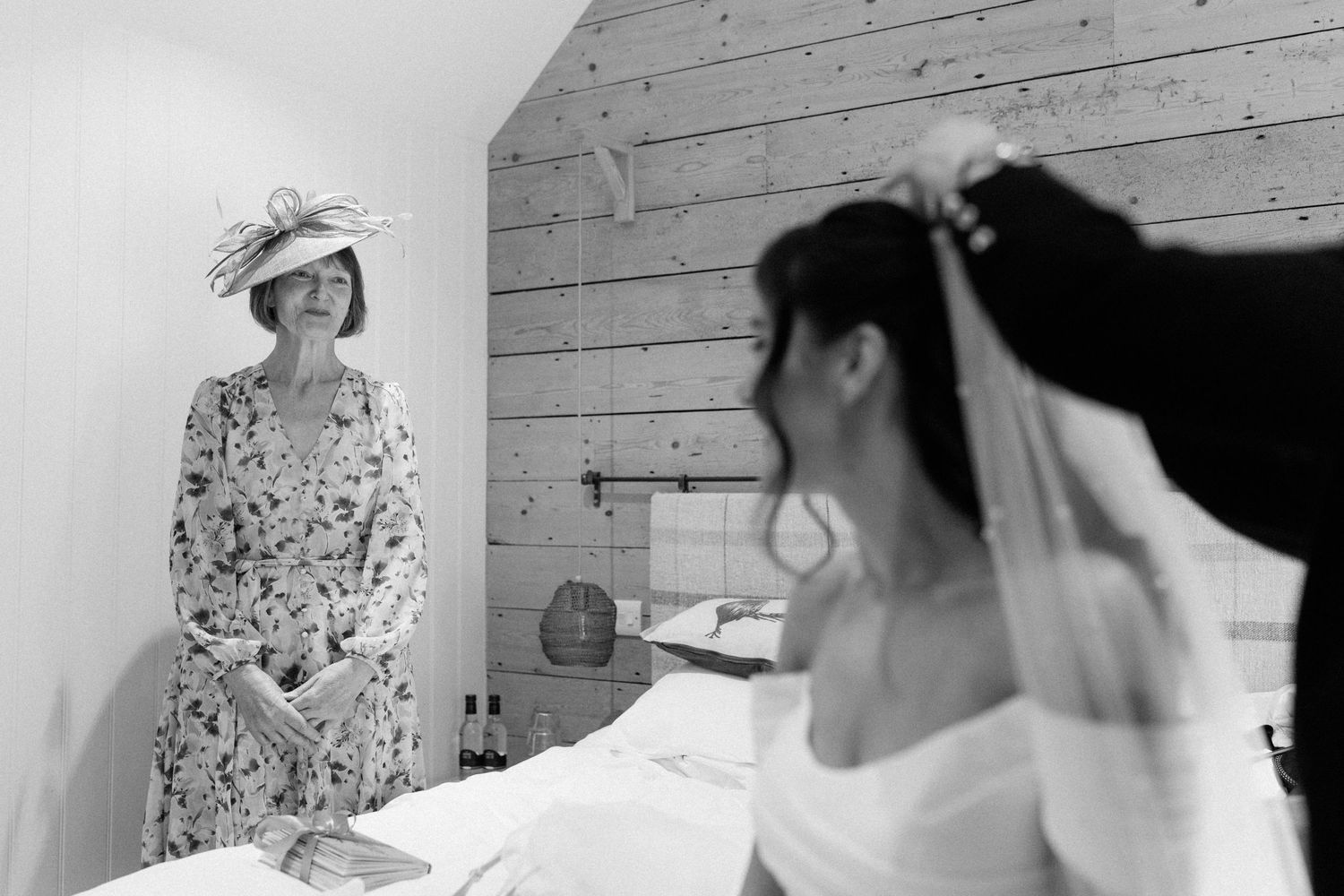Black and white photo of a bride adjusting her veil while a woman in a floral dress and hat watches in a bedroom.