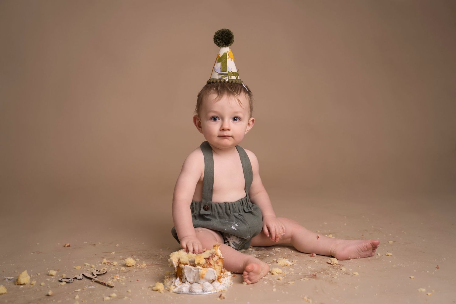 Baby in suspenders and gold party hat smashes first birthday cake on beige studio backdrop.