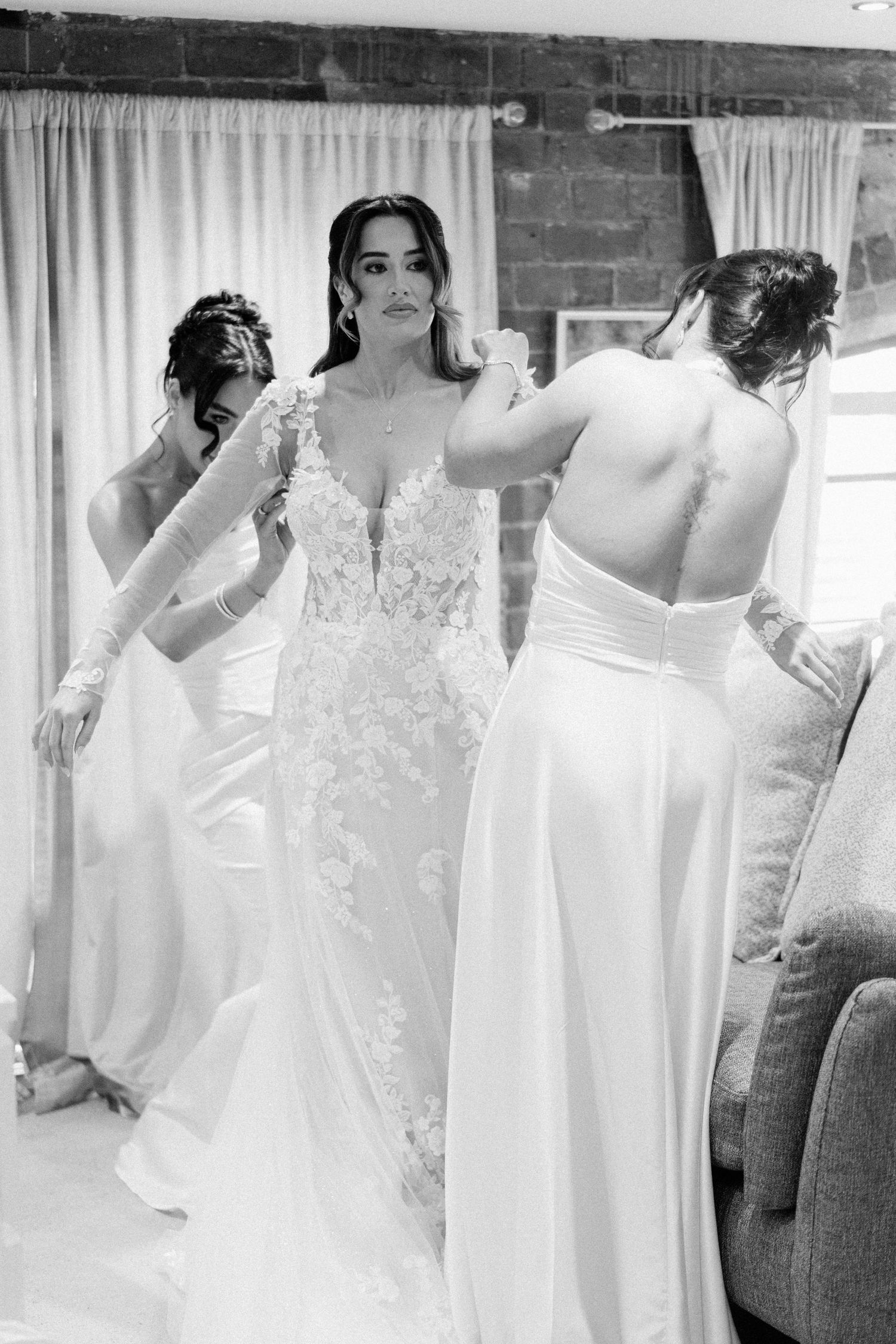 Bride in lace wedding gown being helped by bridesmaids in white dresses during bridal preparations, black and white photo.