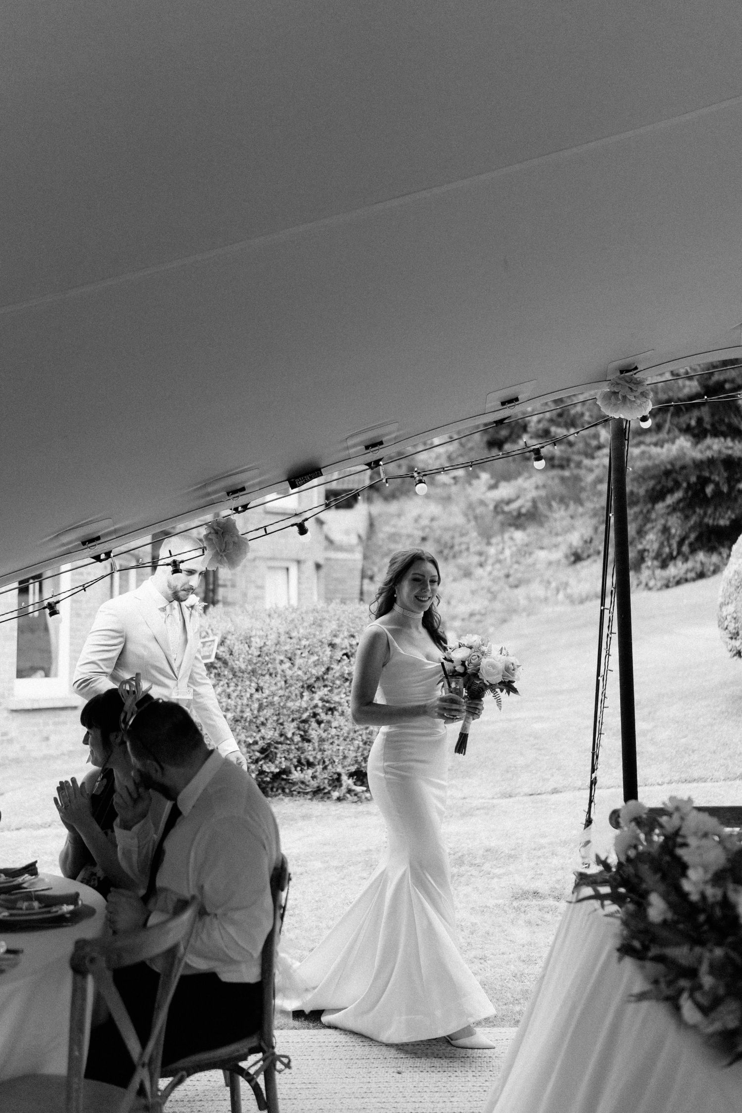 Bride in a fitted white gown walks with bouquet under a tent at an outdoor wedding ceremony.