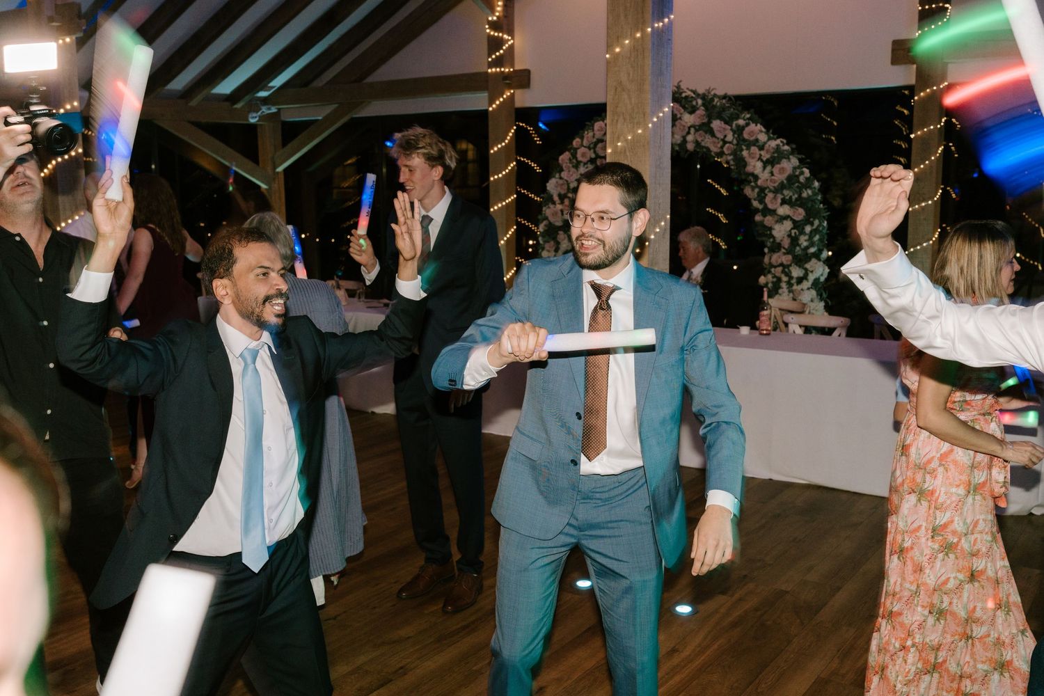 Men in suits dancing with glow sticks at a wedding reception celebration.