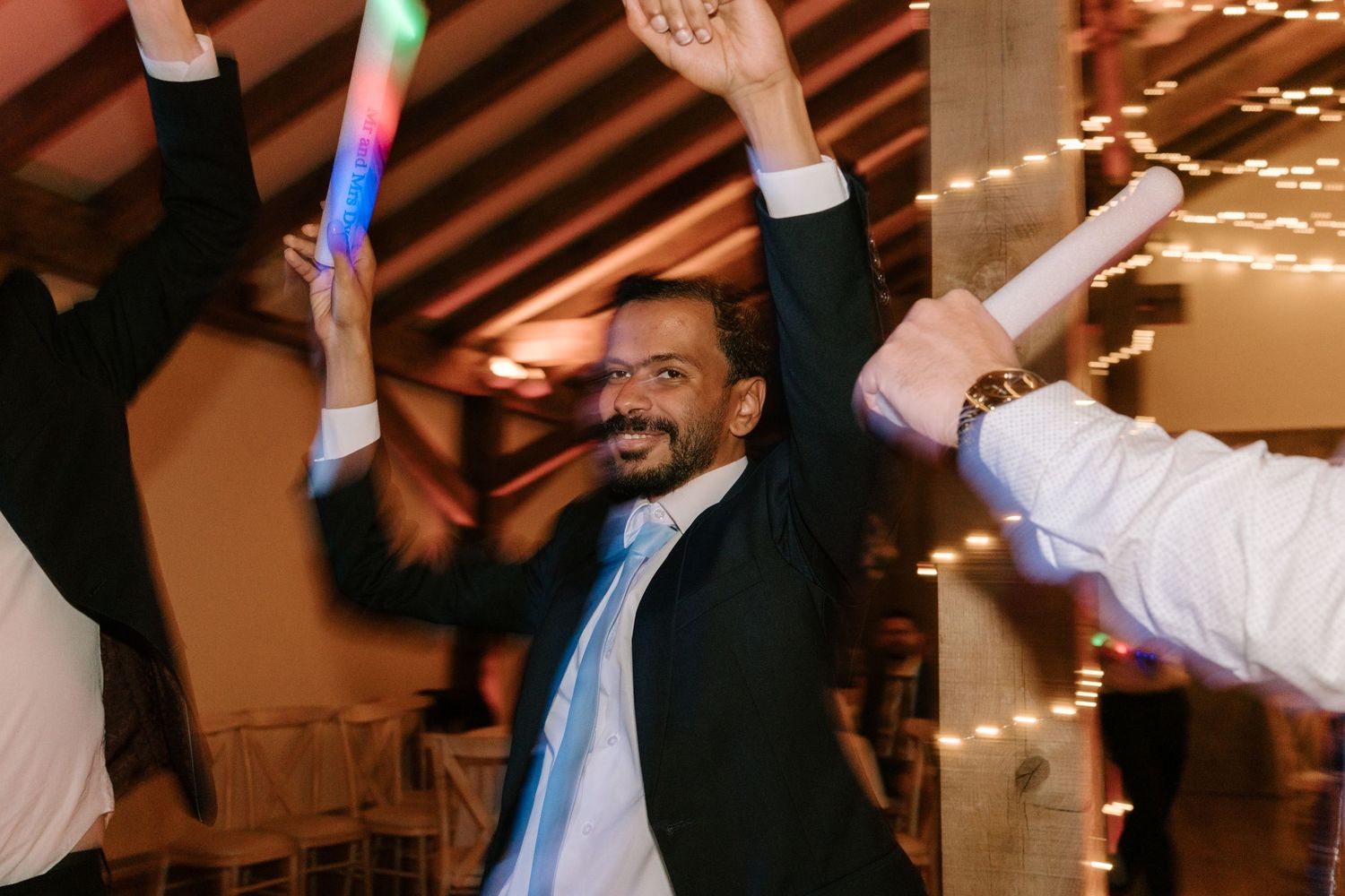 Man in suit dancing joyfully at a wedding reception with colorful glow sticks and string lights in background.