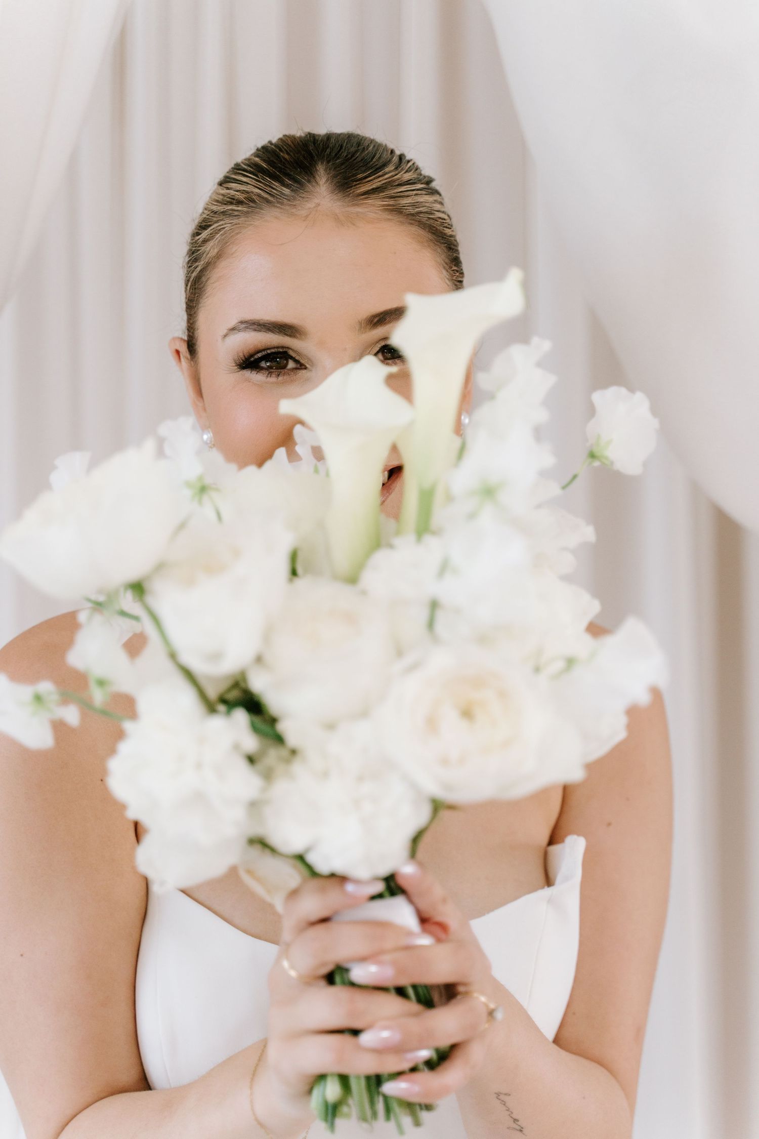 Bride in white dress holding elegant bouquet of white roses and calla lilies, partially covering her face.