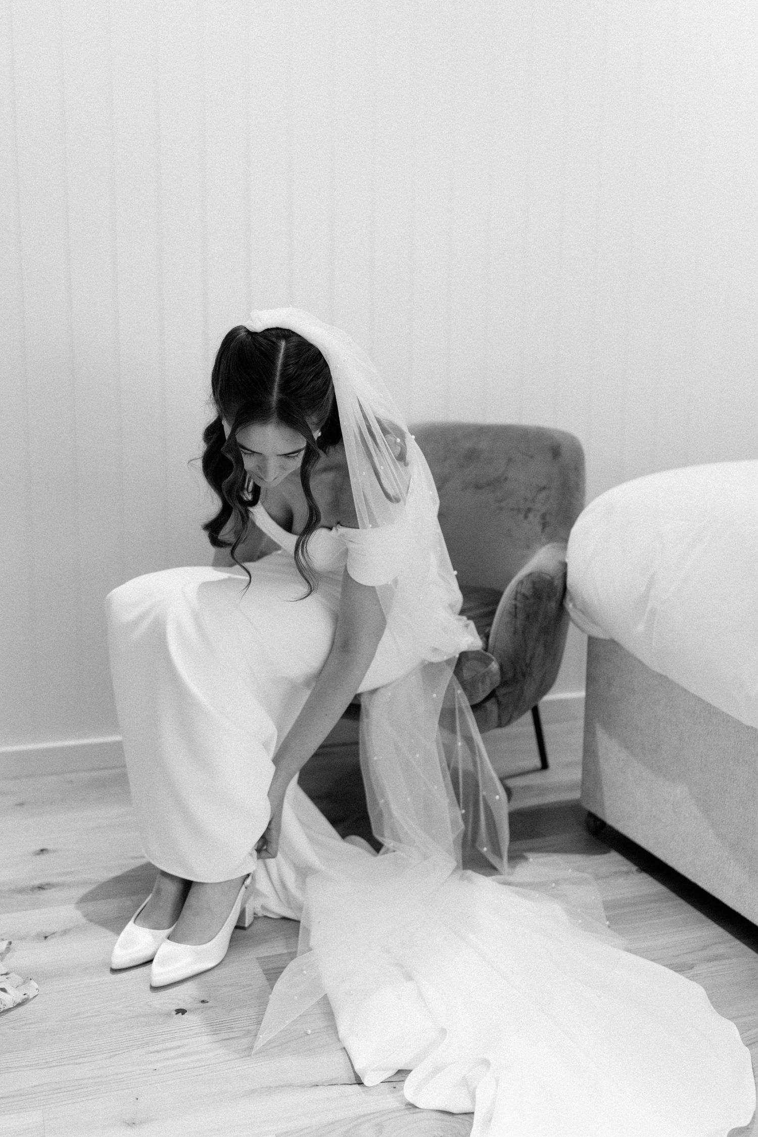 Bride in white gown and veil sitting on floor putting on white heels, black and white wedding photo.