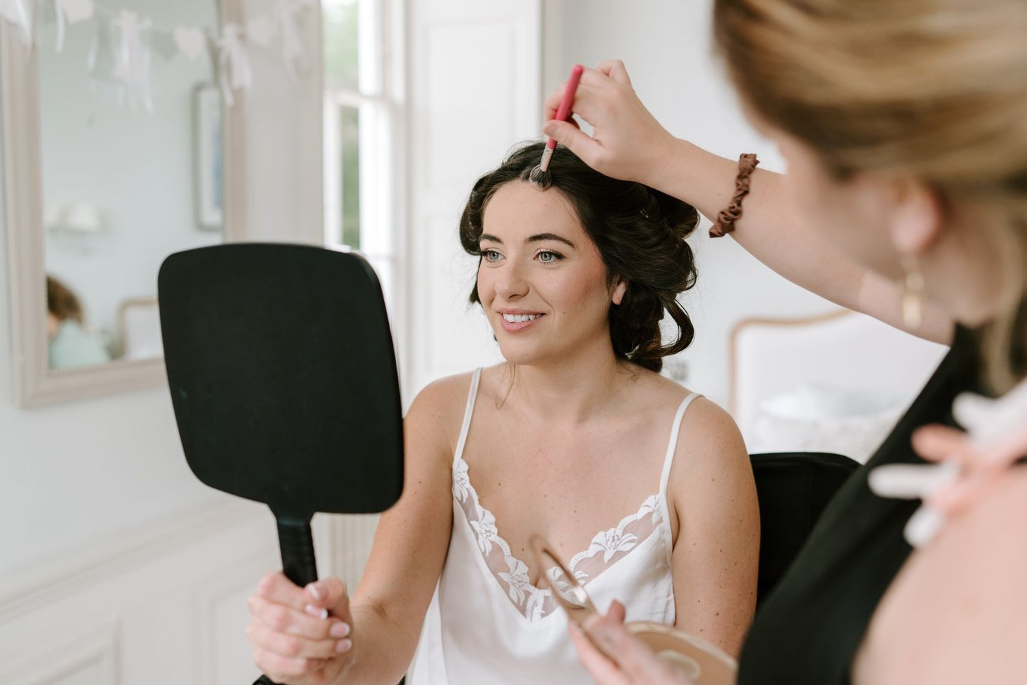Bride smiling while hairstylist works on her updo, holding a hand mirror in a bright, elegant dressing room.