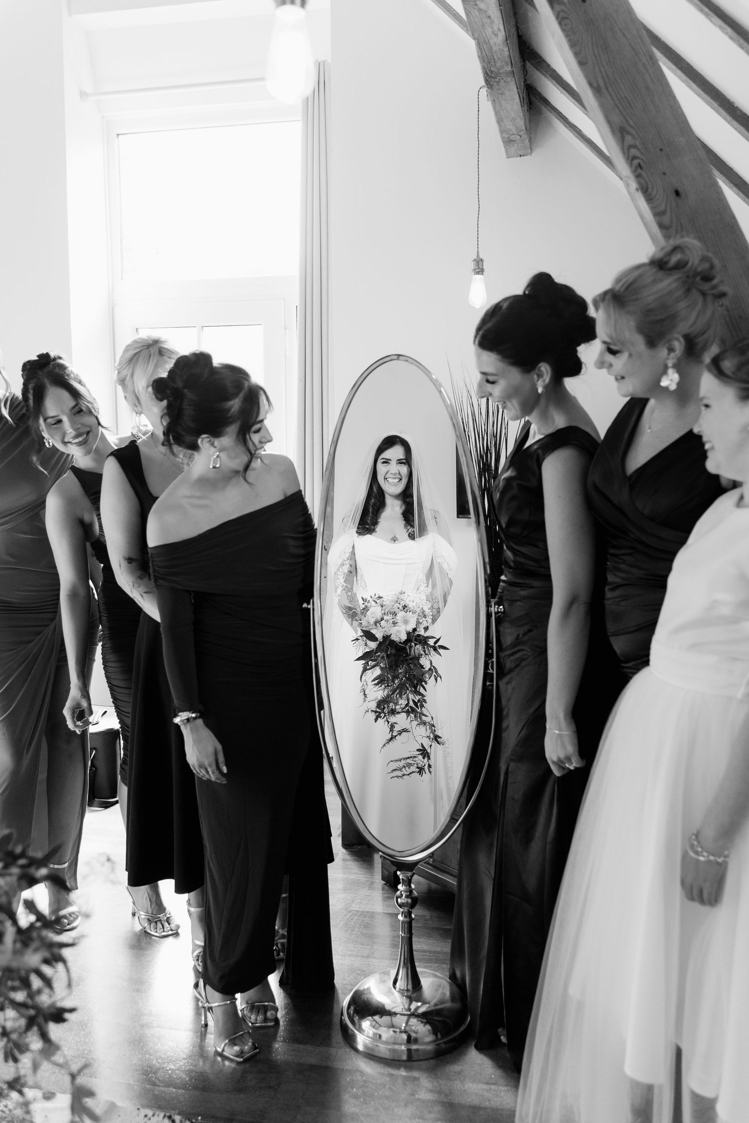 Bride admires her reflection in an oval mirror surrounded by bridesmaids in black dresses during wedding preparations.