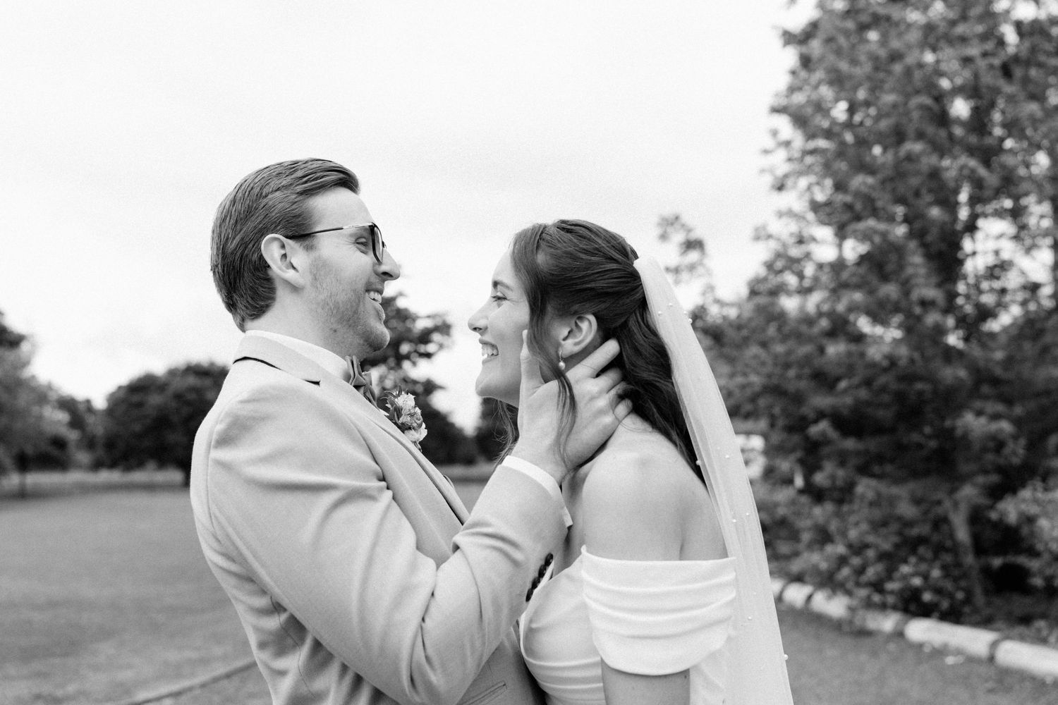 Black and white photo of a groom tenderly holding his bride's face as they share a romantic moment outdoors.