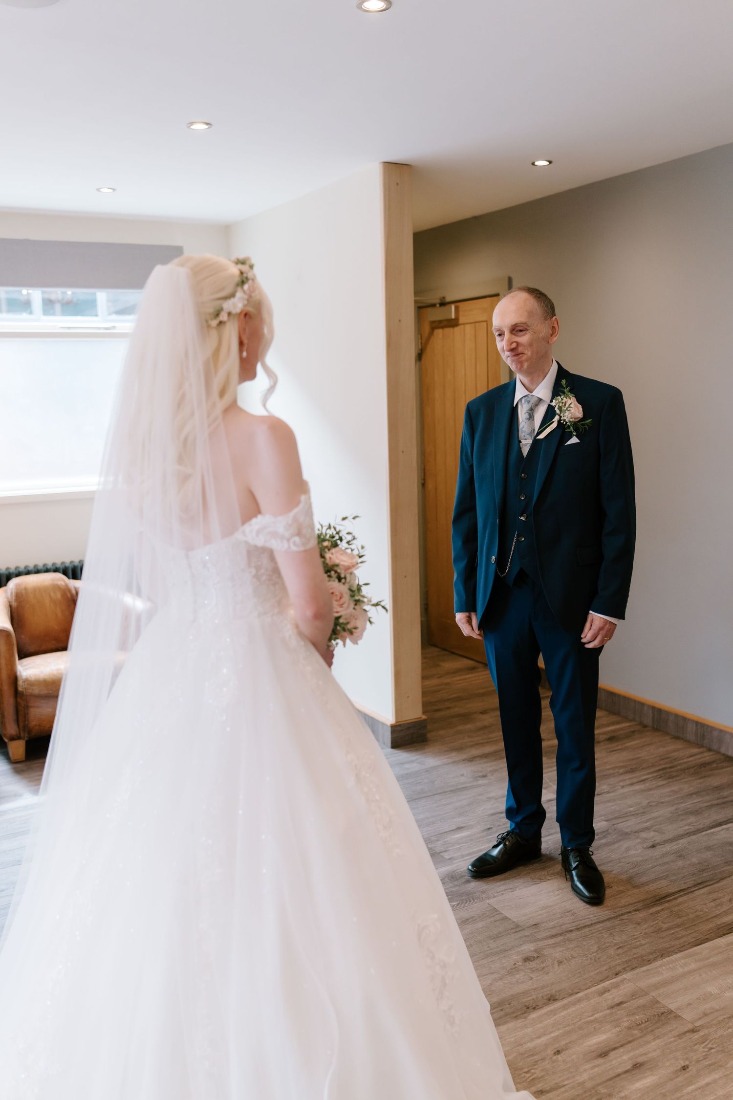 Groom in navy suit smiles at bride in white gown and veil during emotional first look moment indoors.