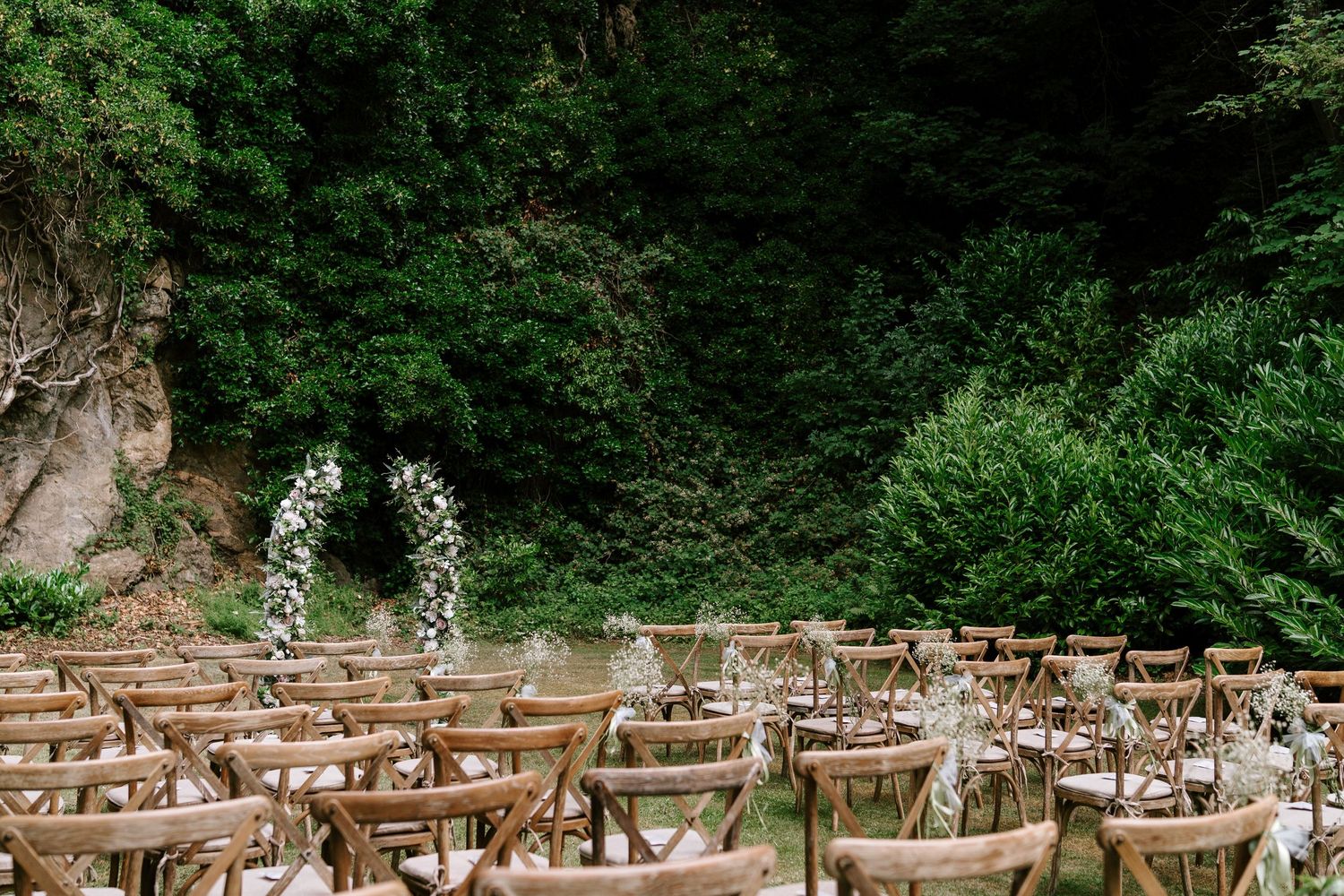 Outdoor wedding ceremony setup with wooden chairs and floral arch against lush green forest backdrop.