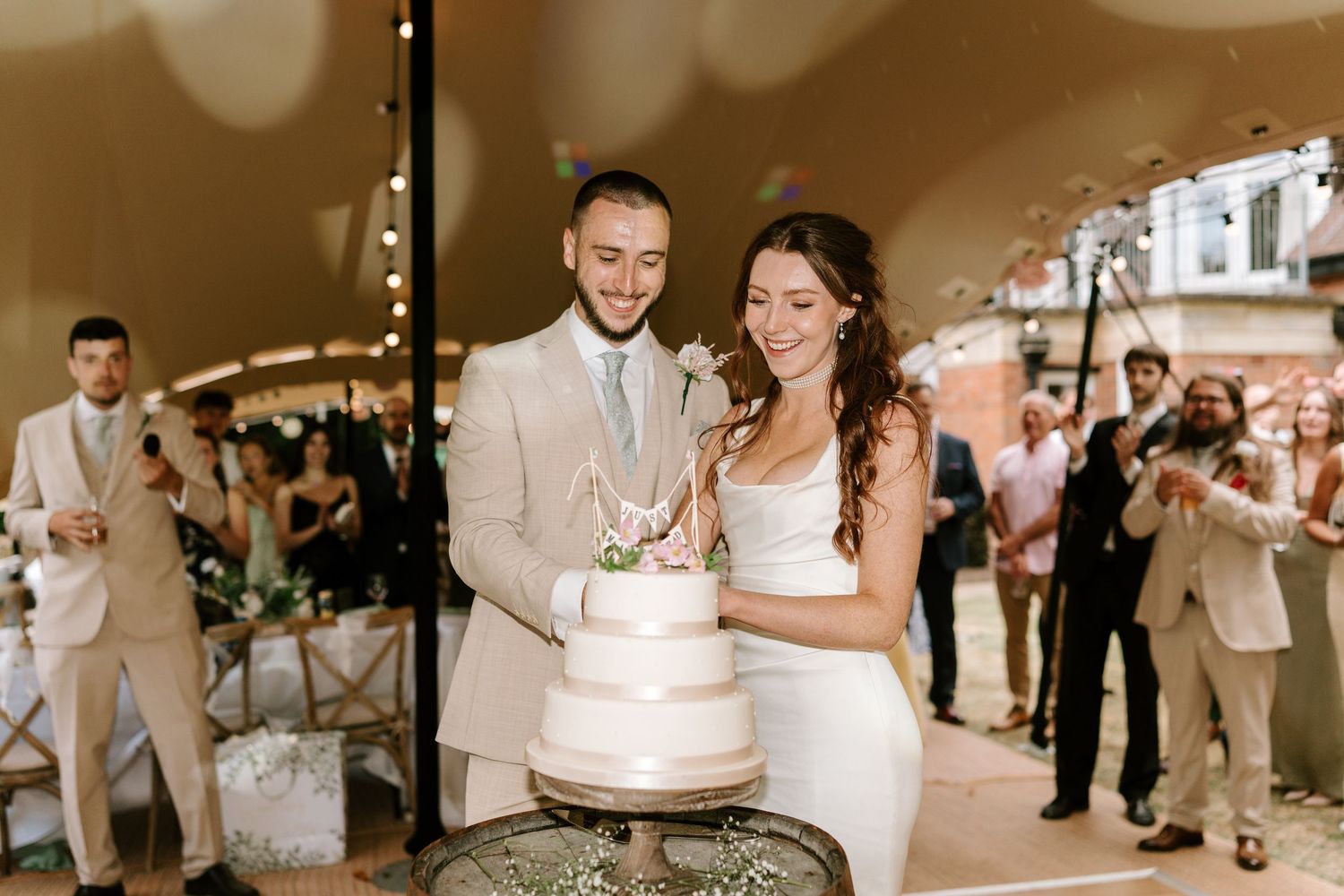 Smiling newlyweds cutting three-tier white wedding cake at elegant indoor reception surrounded by guests.