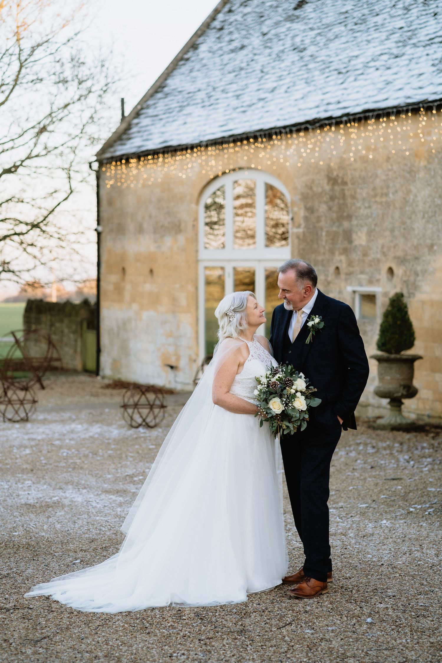 Bride and groom share a romantic moment outside a rustic stone venue decorated with fairy lights on their winter wedding day.