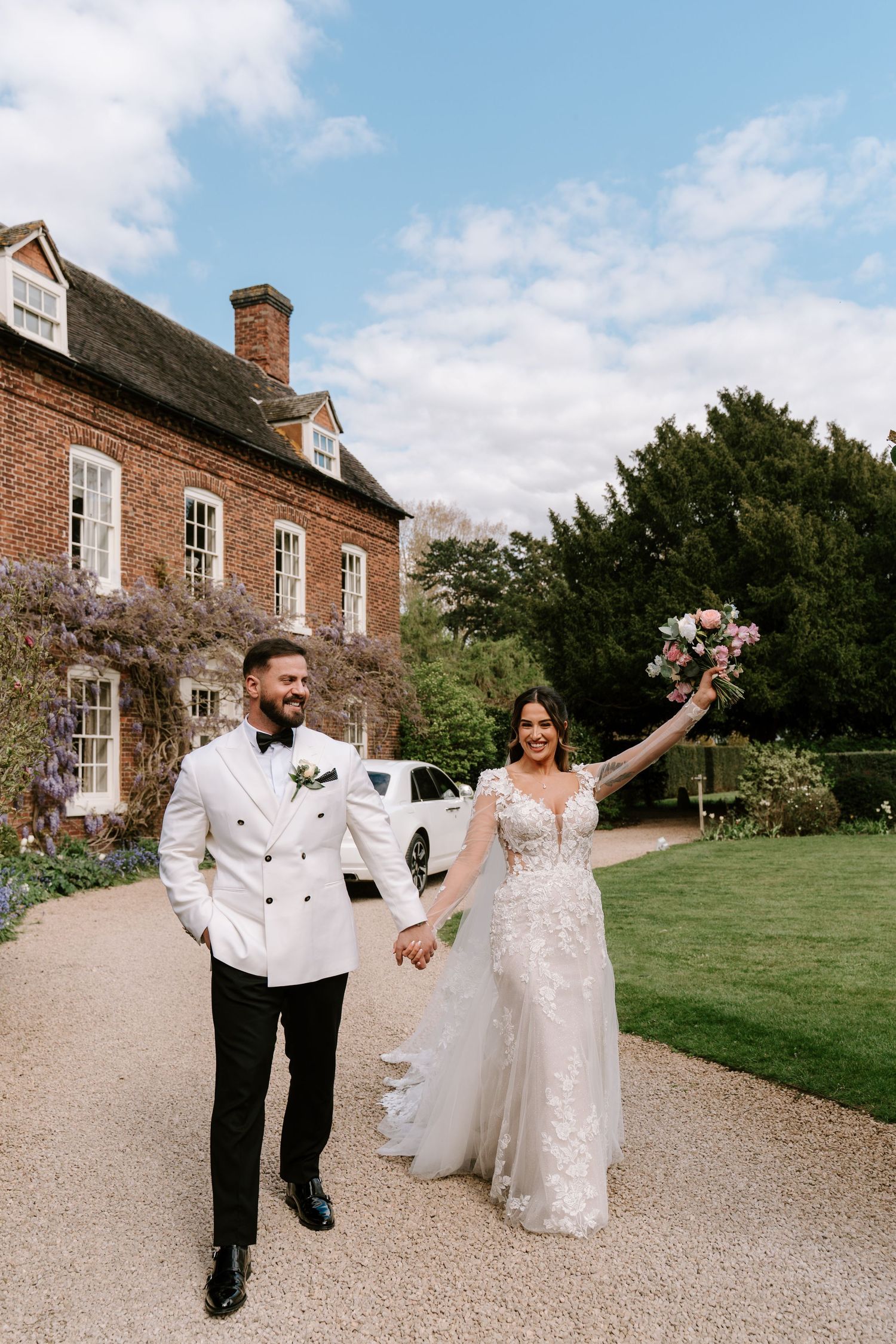 Bride and groom walk hand-in-hand outside a brick manor, bride in lace gown raising bouquet, groom in white tuxedo.