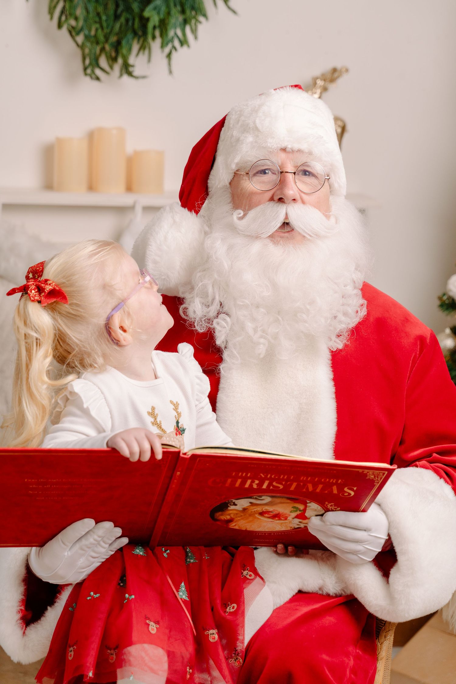 Santa Claus reading a Christmas book with a young blonde girl in a festive holiday setting.