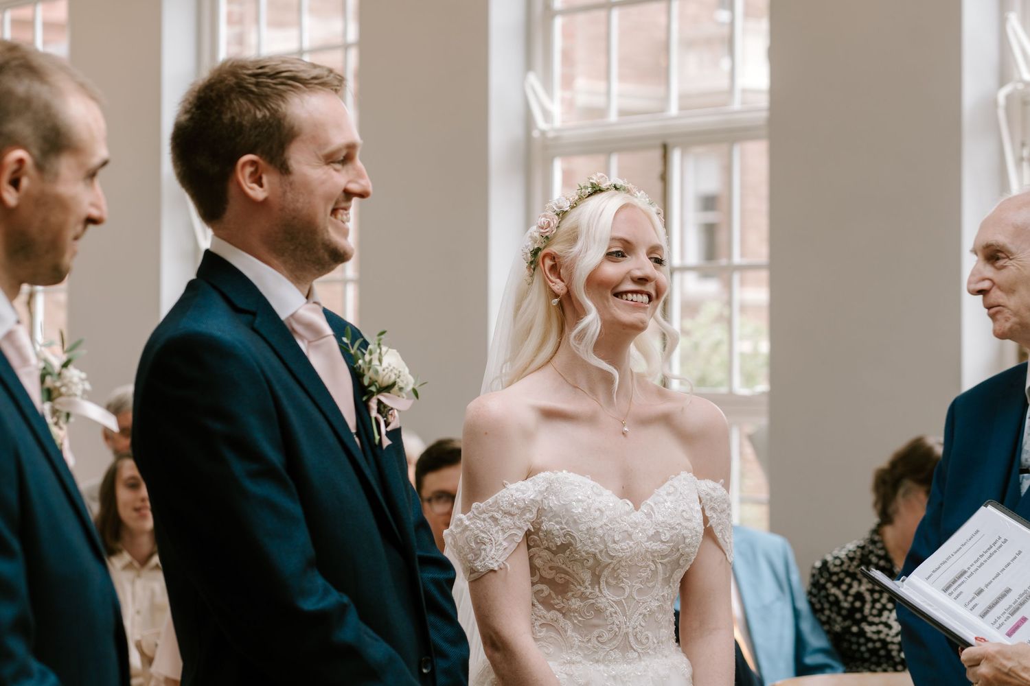 Bride and groom smiling joyfully during wedding ceremony, bride wearing off-shoulder lace gown with floral hair accessory.