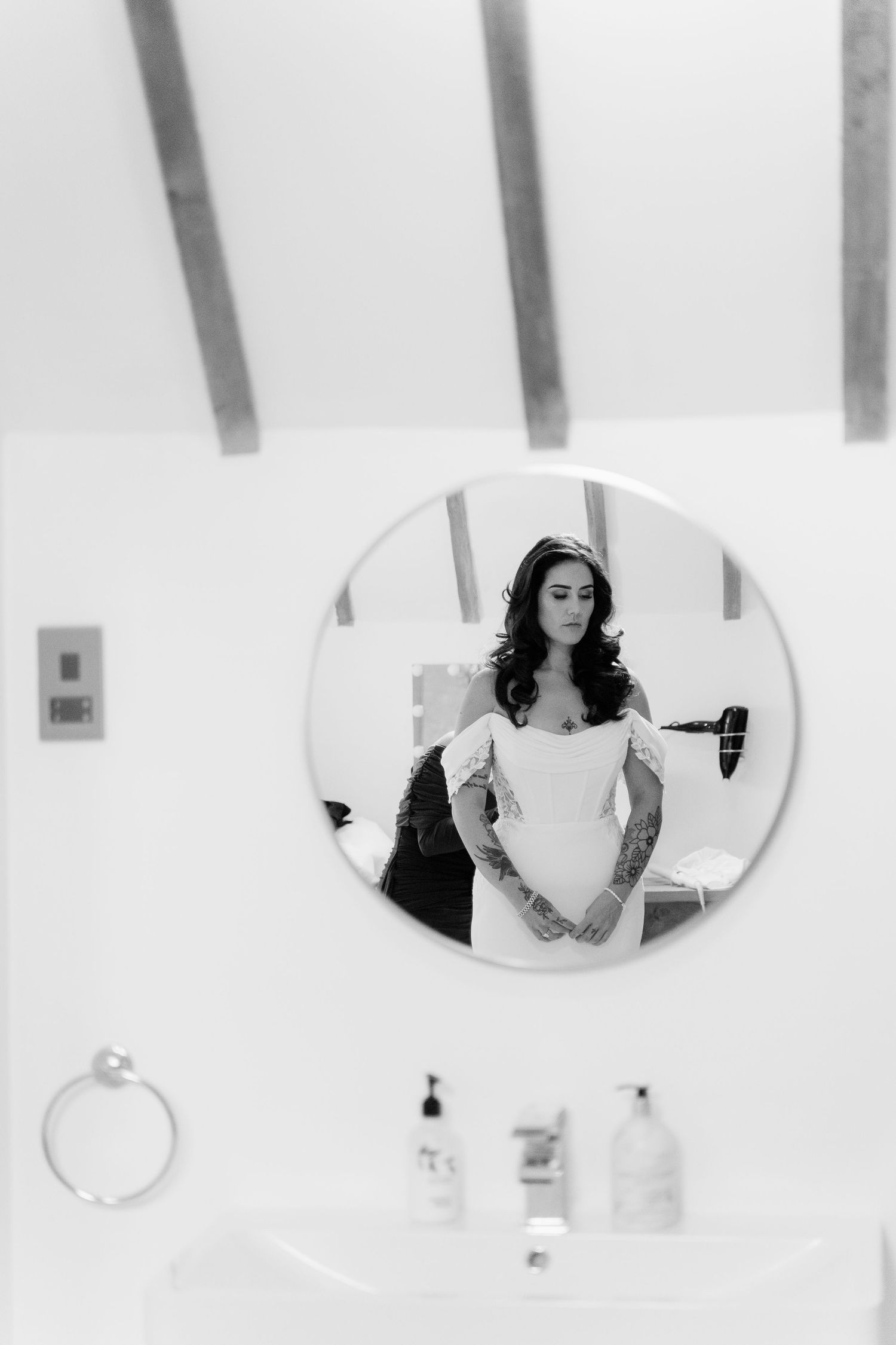 Black and white photo of tattooed bride in white dress reflected in round bathroom mirror on wedding day.