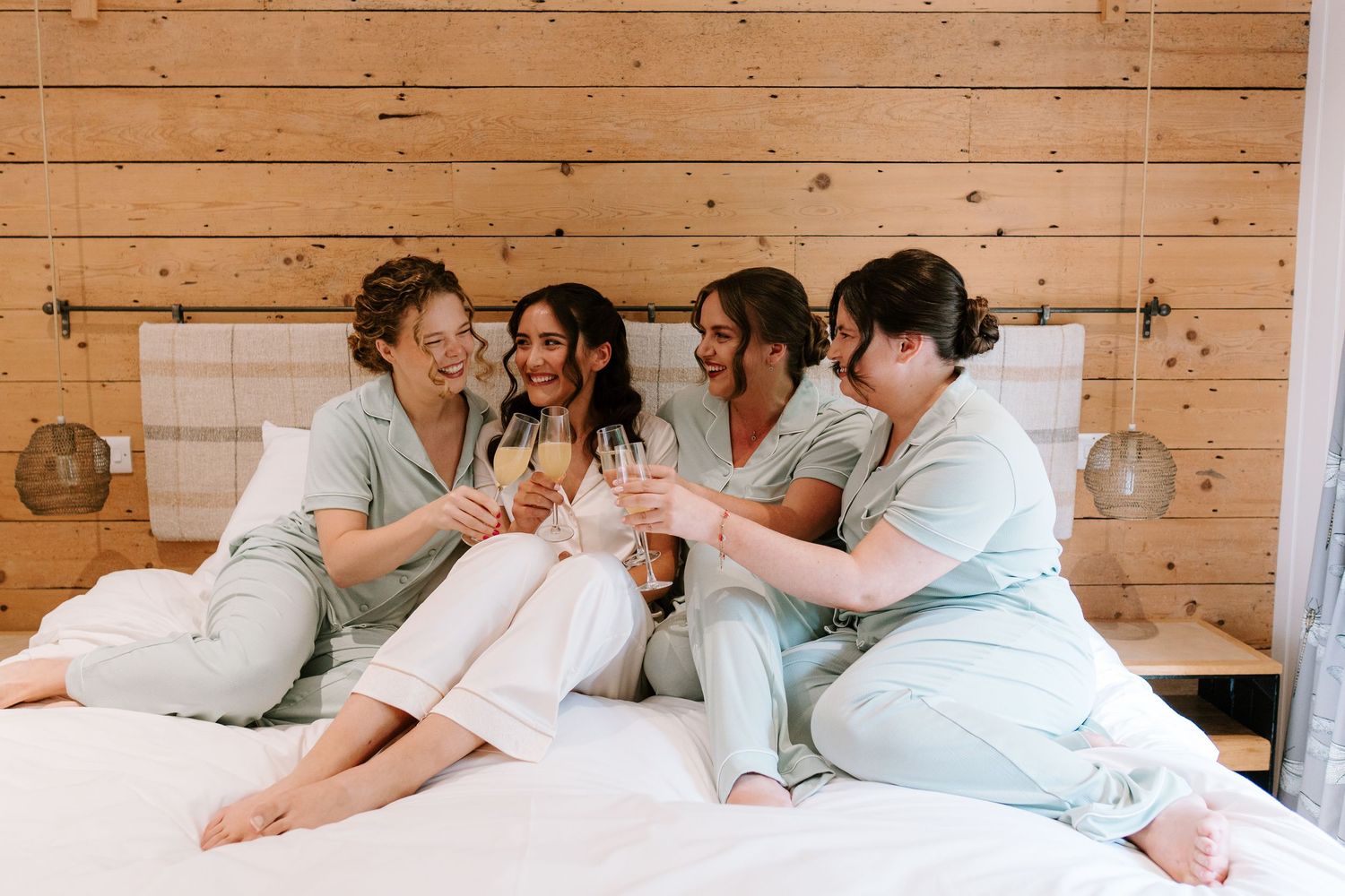 Bridesmaids in matching pajamas toasting with champagne on a white bed, laughing together before the wedding.