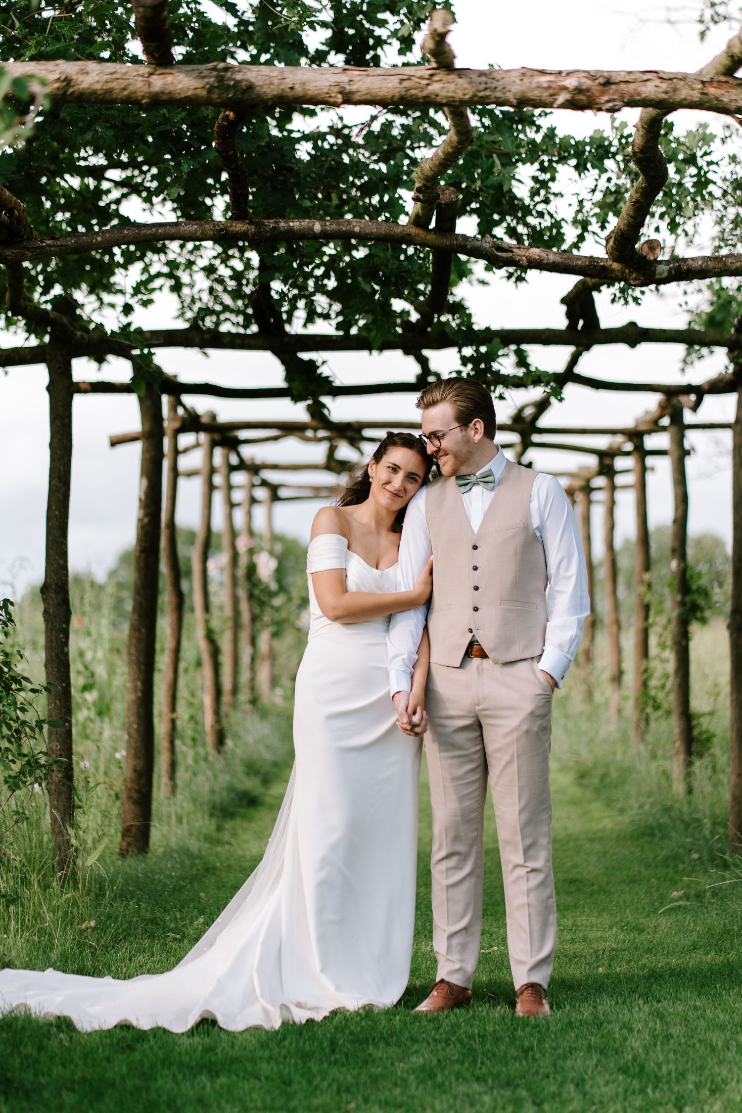 Bride and groom holding hands under a rustic wooden pergola surrounded by lush greenery at an outdoor vineyard wedding.