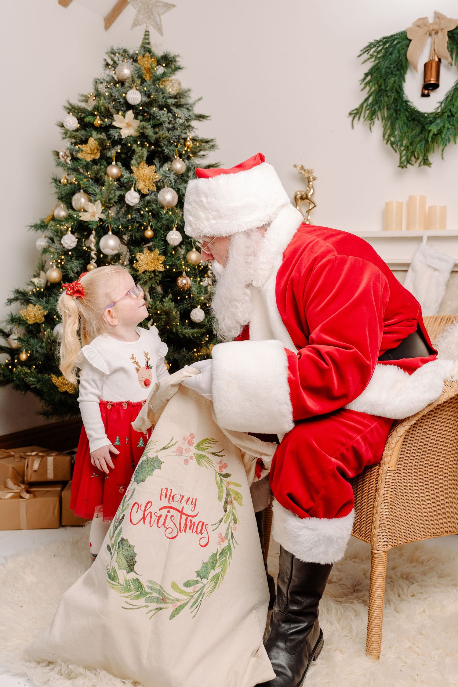 Santa Claus showing a Merry Christmas gift sack to an excited little blonde girl near a decorated Christmas tree.