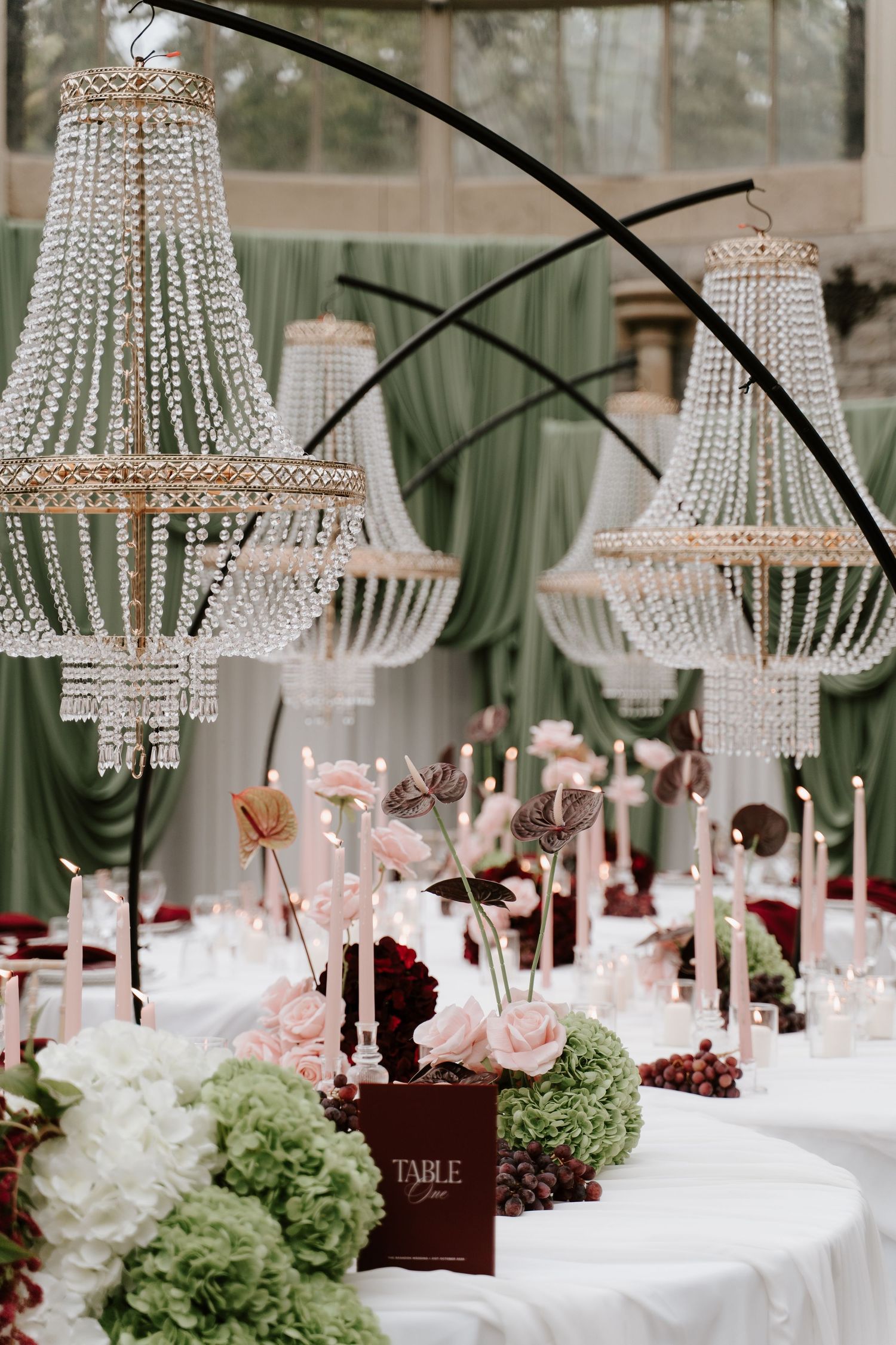 Elegant wedding reception table with crystal chandeliers, pink roses, green hydrangeas, and candles under a draped green ceiling.