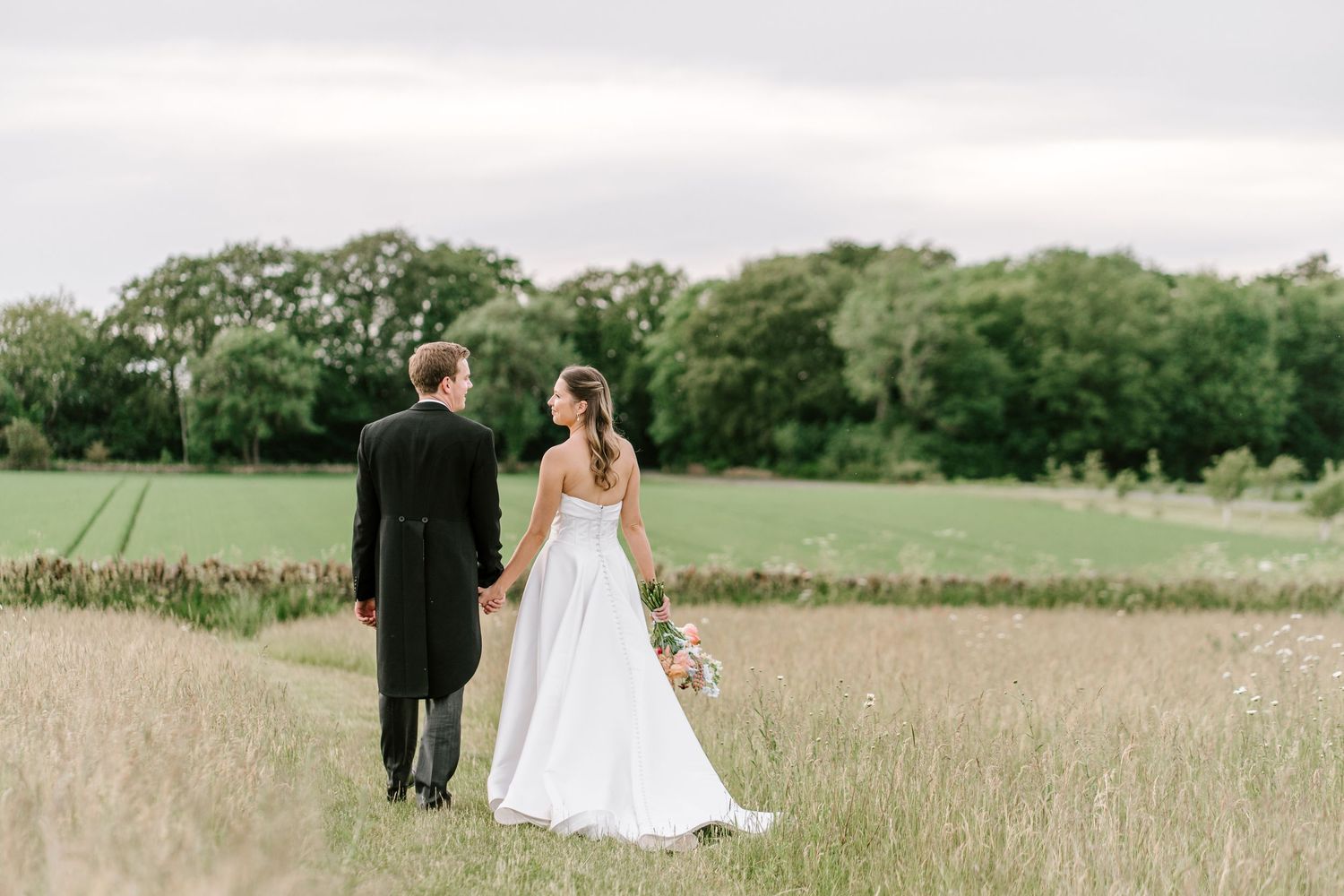 Bride and groom walk hand-in-hand through a lush green field on their wedding day.