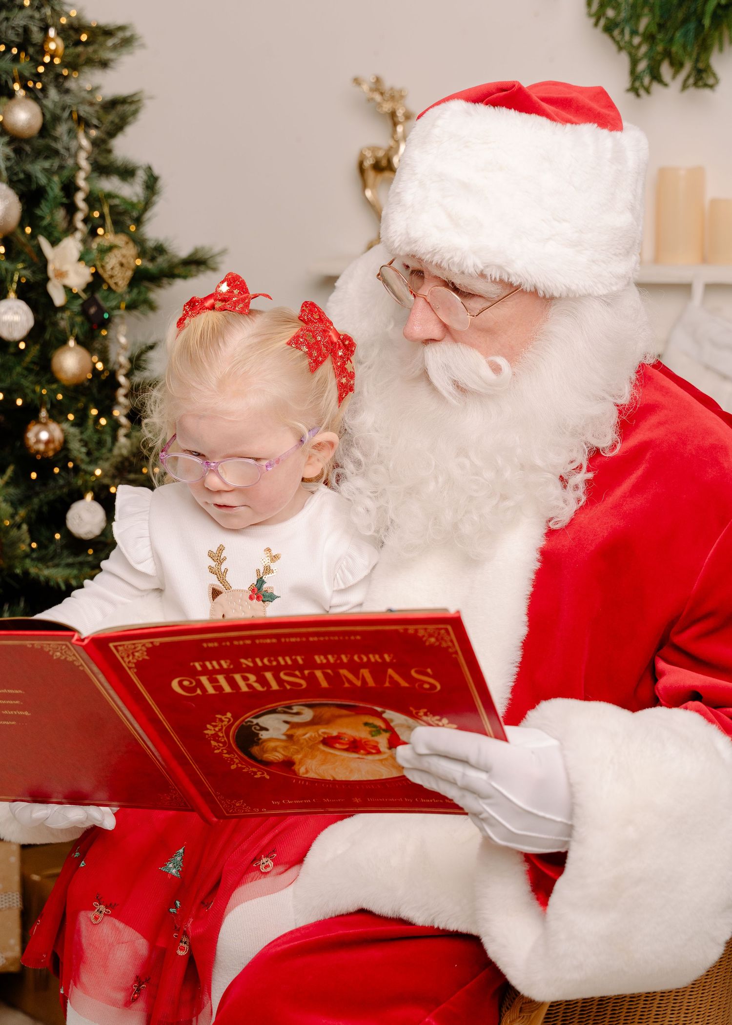 Santa Claus reads The Night Before Christmas book to a little girl wearing a reindeer outfit and red bow.
