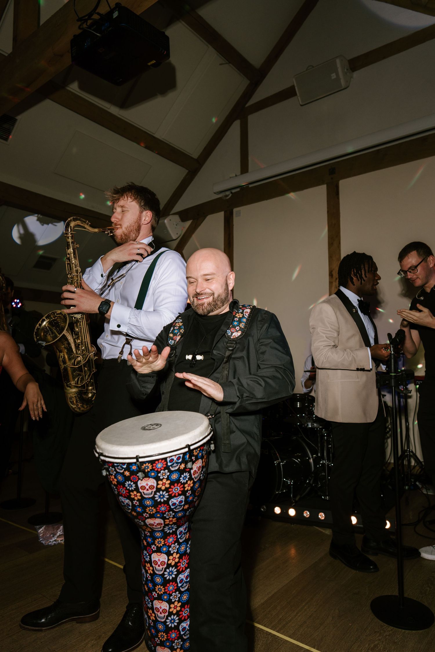Bald drummer plays colorful skull bongo drums beside saxophonist at lively wedding reception event.