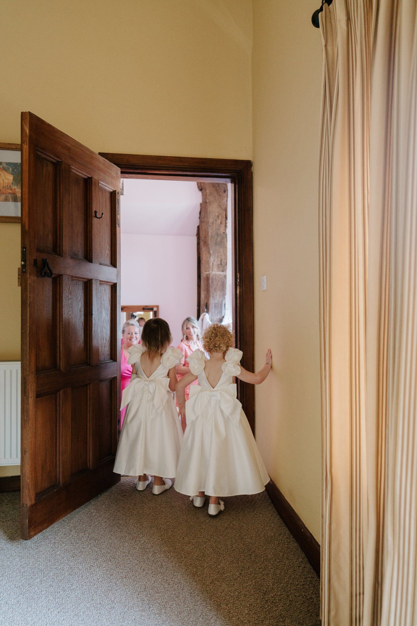 Two flower girls in white dresses walk through a wooden doorway during a wedding ceremony preparation.