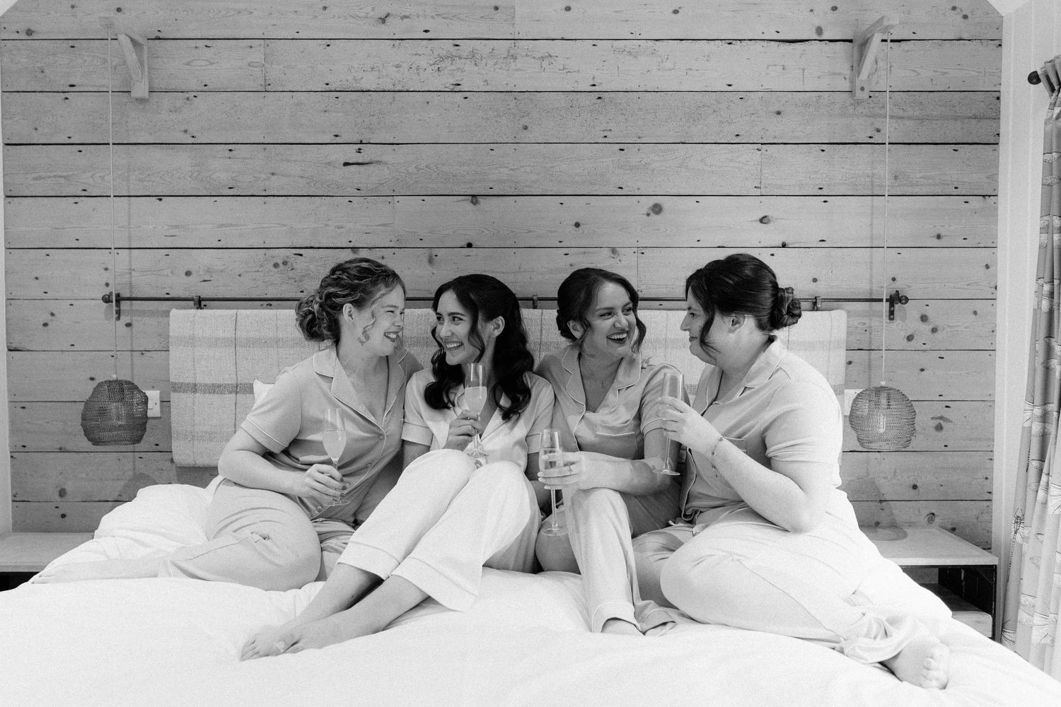 Four bridesmaids in matching pajamas laughing and toasting with champagne on a bed in a black and white wedding photo.