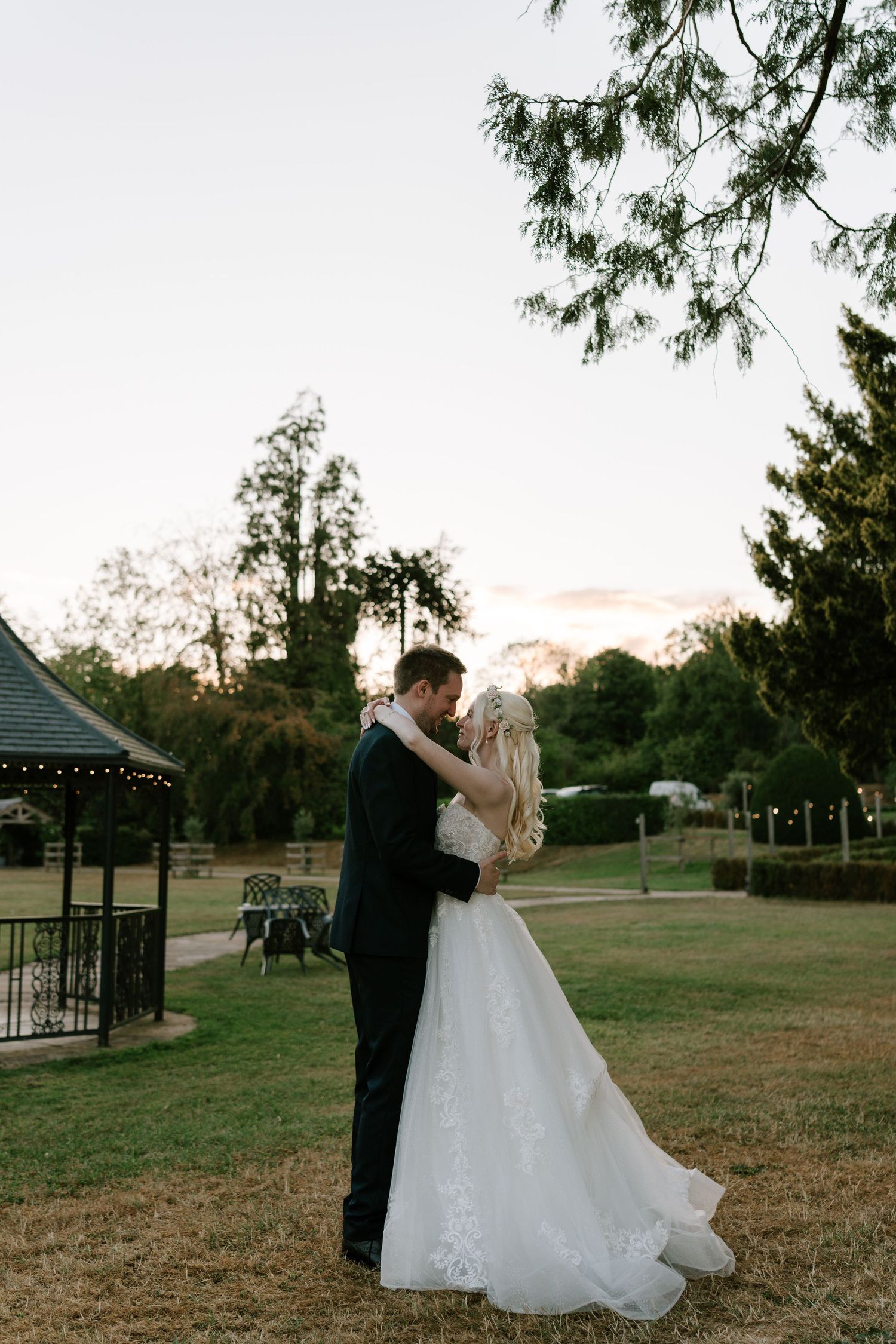 Bride and groom share a romantic embrace at sunset in a lush garden, bride wearing elegant white lace gown.